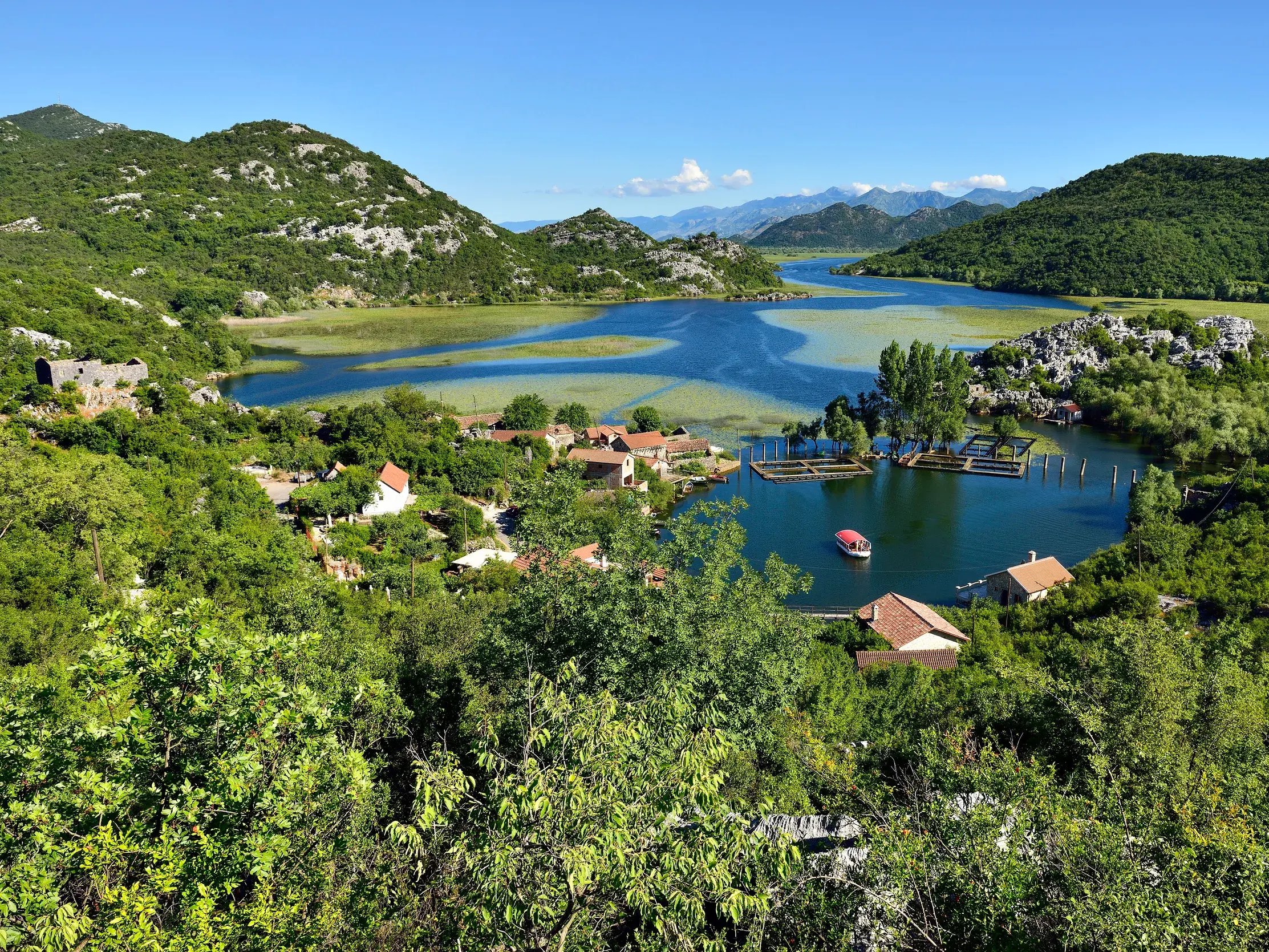 Panoramablick von einer Wanderung auf den blauen Skadar-See in Montenegro, umgeben von grünen Bergen und einem kleinen Dorf am Ufer.