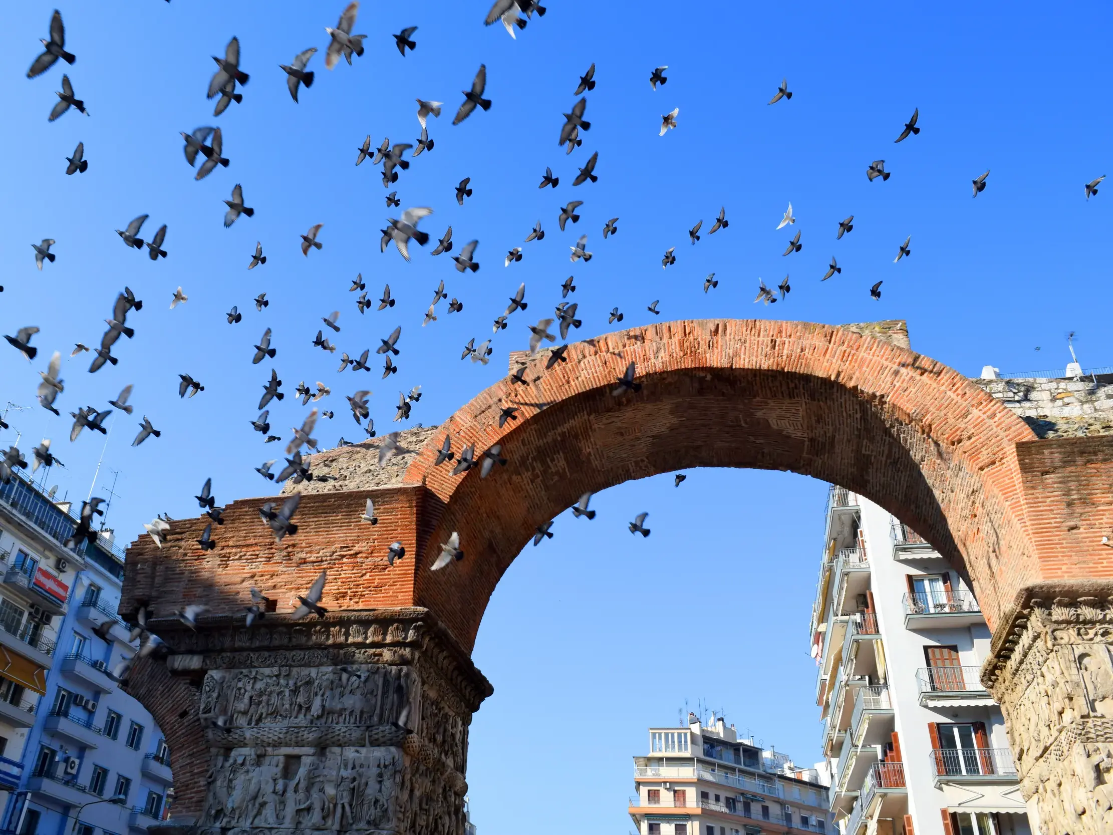 Ein großer Schwarm Tauben fliegt über einen antiken Torbogen aus Backstein in einer griechischen Stadt unter blauem Himmel.