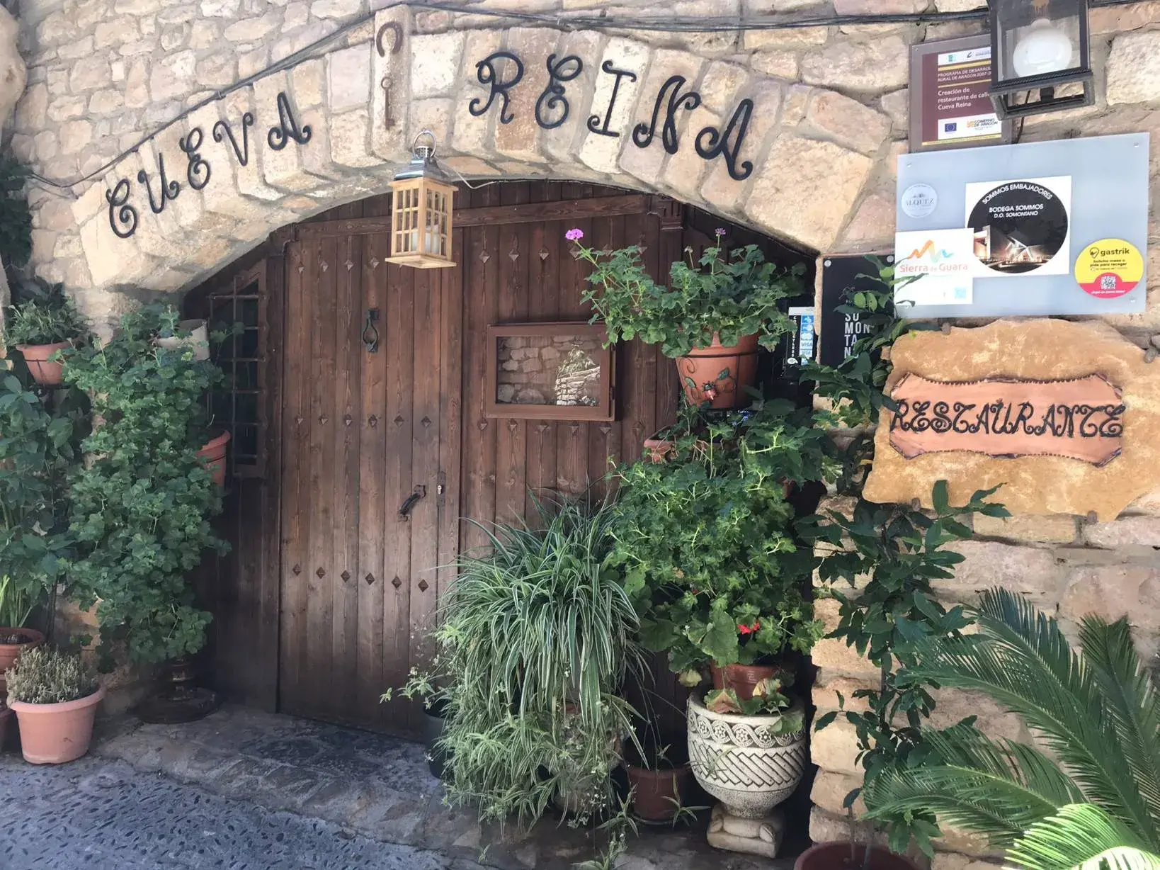 Der charmante Eingang des Restaurants Cueva Reina in Alquézar mit rustikalem Steinbogen, Holztür und üppigen Grünpflanzen.