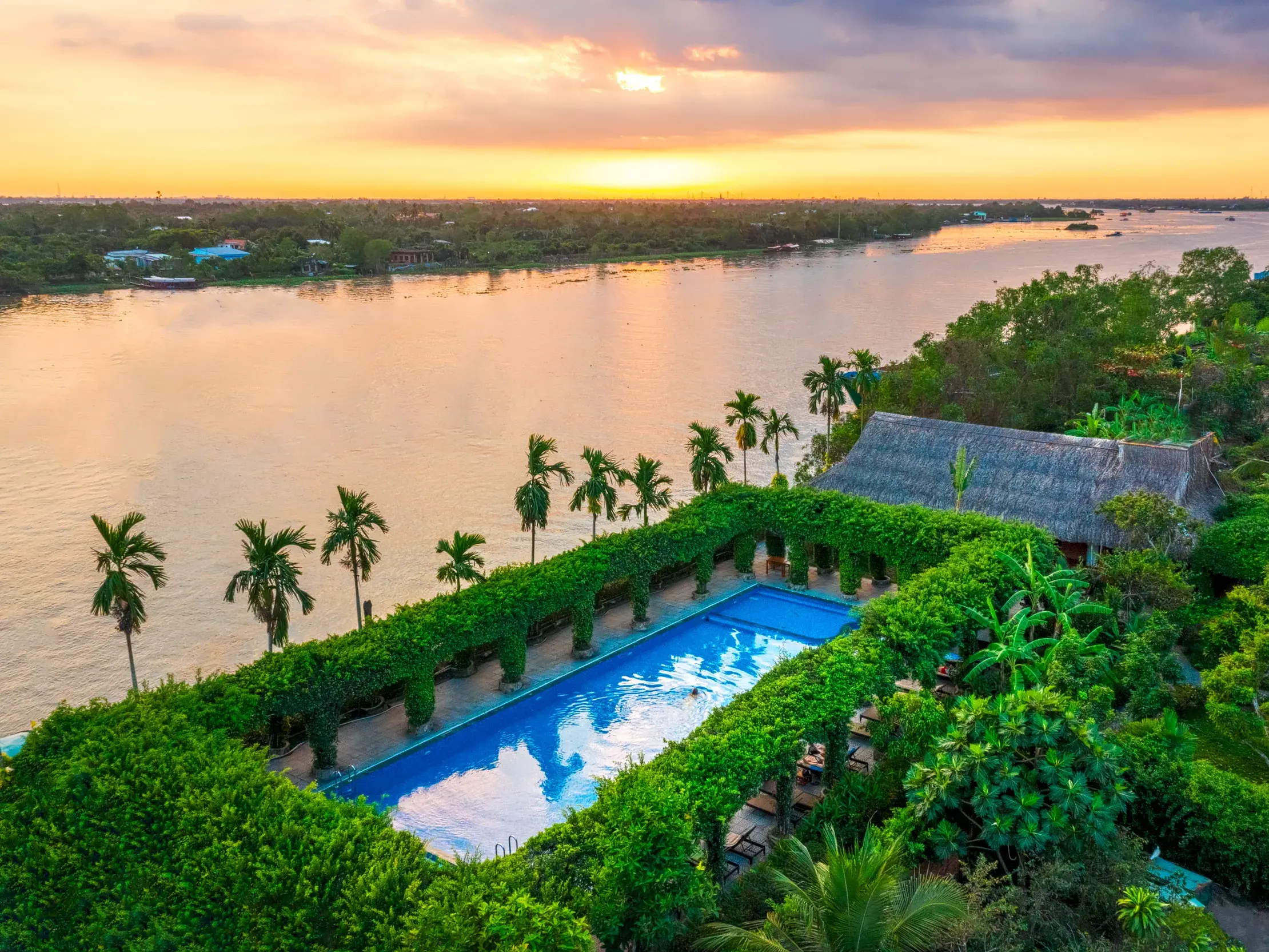 Blick vom Pool der Mekong Lodge mit tropischem Garten auf den breiten Mekong Fluss bei malerischem Sonnenuntergang in Vietnam.