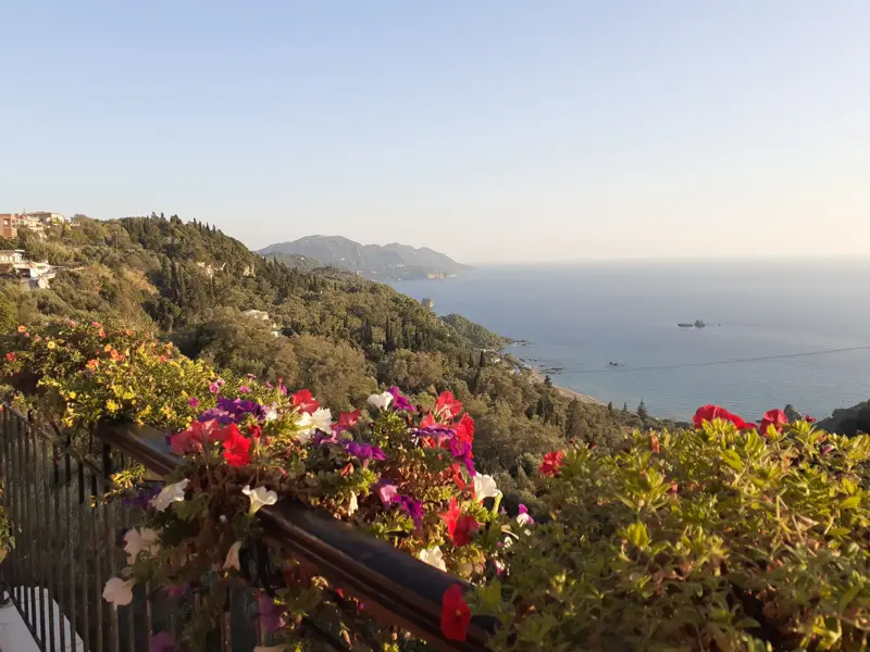 Ausblick von einer Terrasse mit bunten Blumen auf die bewaldete Küstenlandschaft und das Ionische Meer auf Korfu.