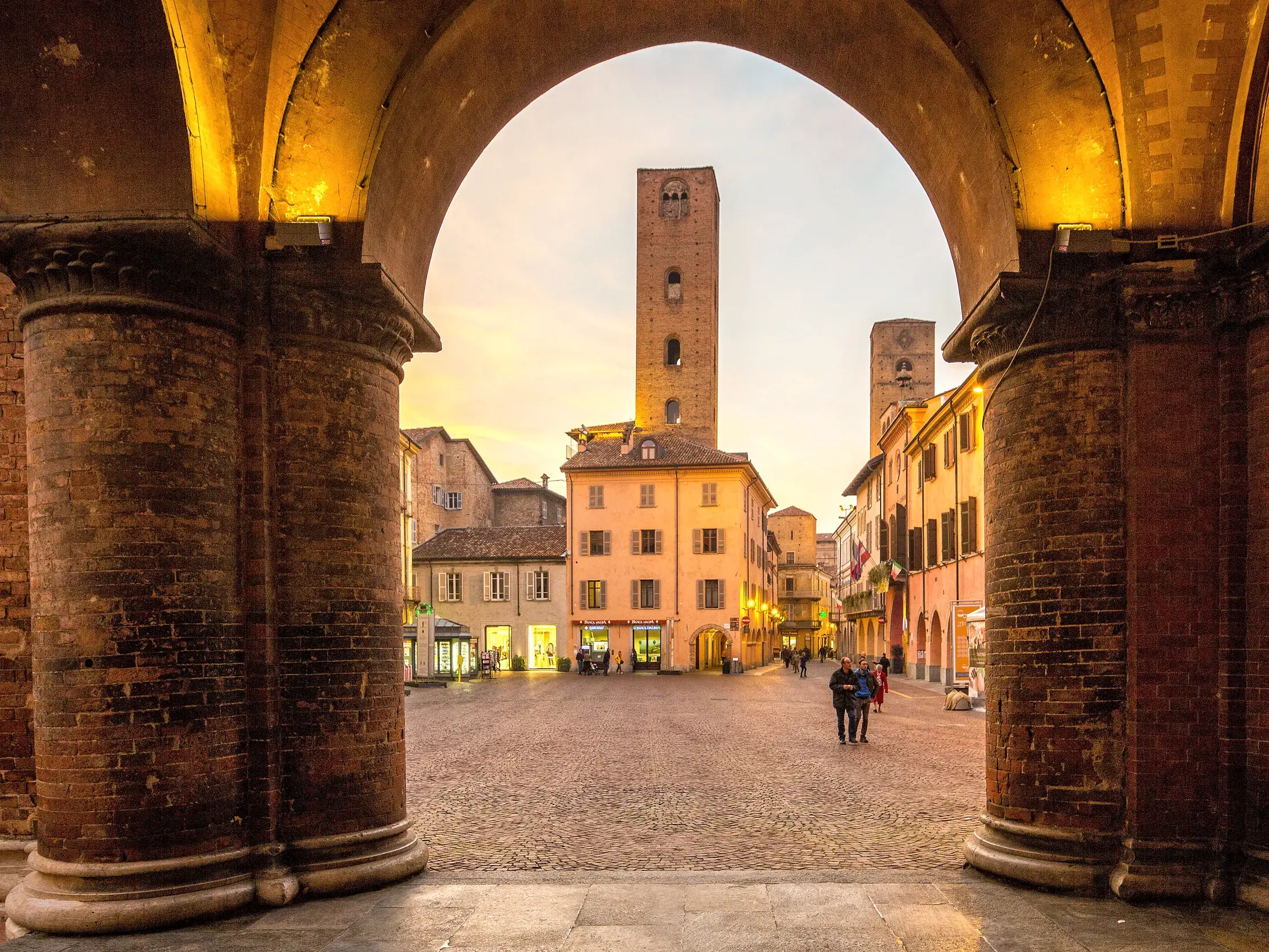Abendlicher Blick durch einen Backstein-Torbogen auf die historische Piazza mit den mittelalterlichen Geschlechtertürmen von Alba.