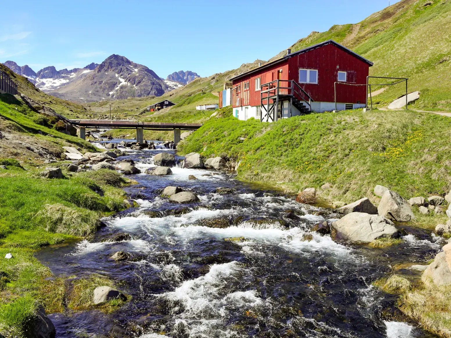 Rotes Holzhaus an einem Gebirgsbach im grünen Blumental bei Tasiilaq, im Hintergrund die Berge Ostgrönlands.