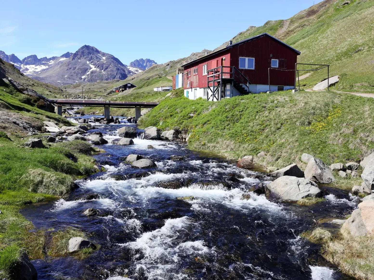 Rauschender Gebirgsbach im grünen Blumental bei Tasiilaq mit einem roten Haus und arktischen Bergen im Hintergrund.