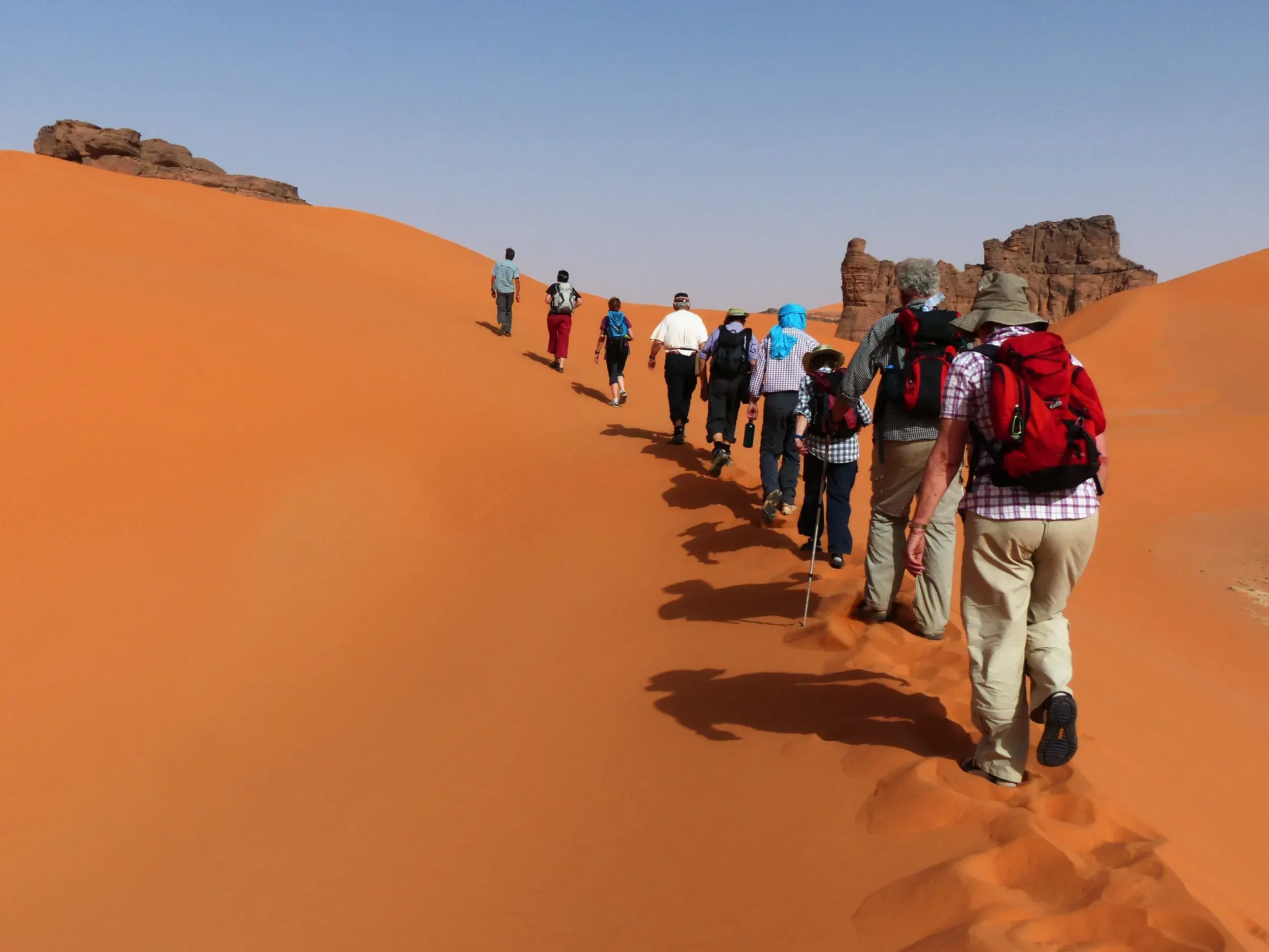 Eine Trekkinggruppe wandert in einer Reihe eine hohe, orangefarbene Sanddüne im Erg Admer in der algerischen Sahara hinauf.