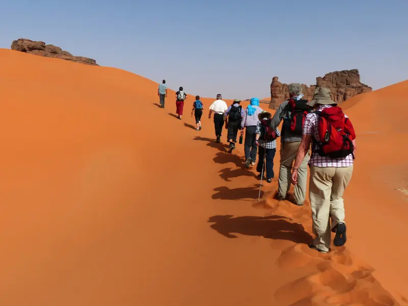Eine Trekkinggruppe wandert in einer Reihe eine hohe, orangefarbene Sanddüne im Erg Admer in der algerischen Sahara hinauf.