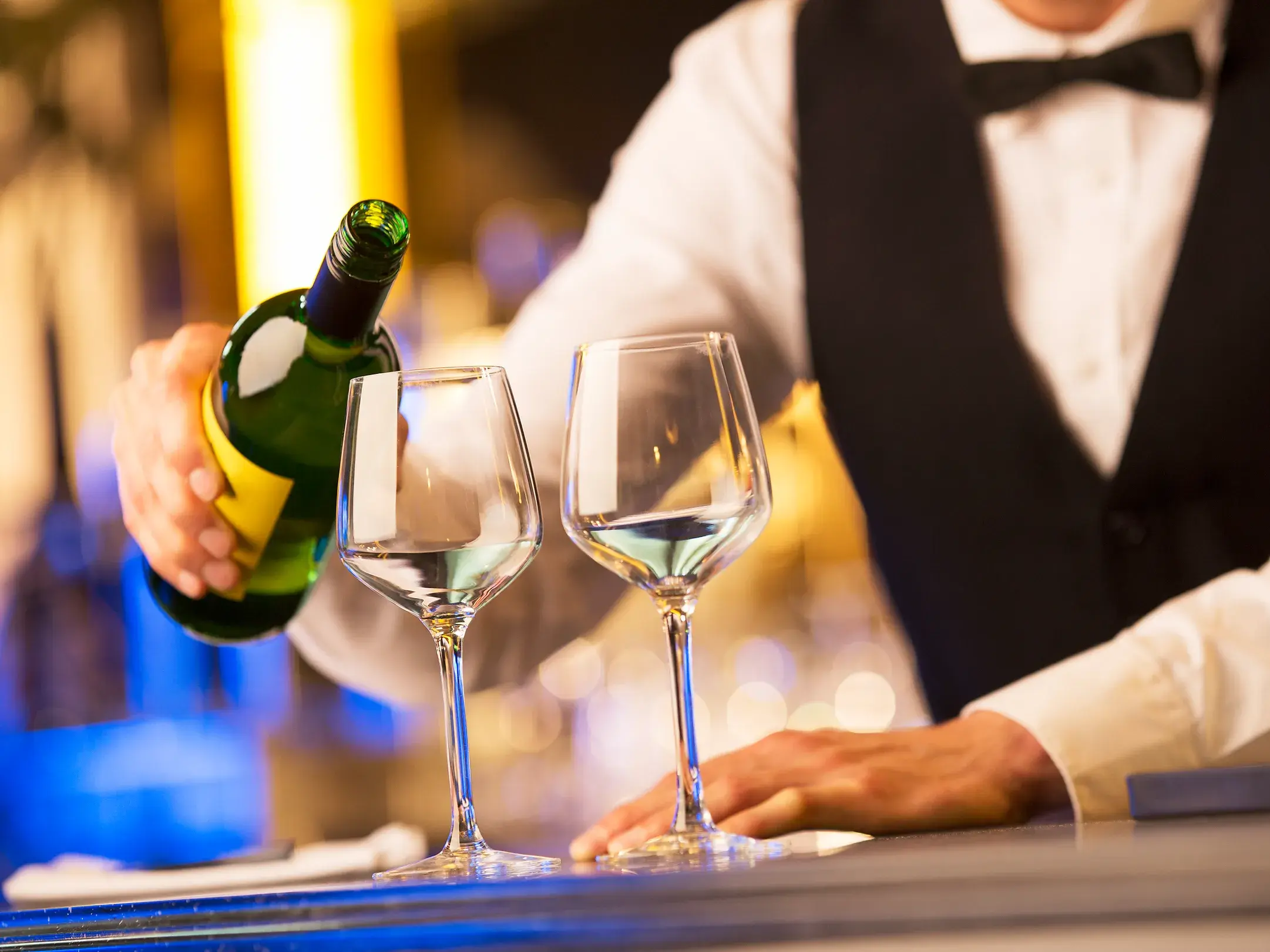 Elegant gekleideter Kellner schenkt an einer Bartheke Weißwein in zwei stilvolle Gläser ein.