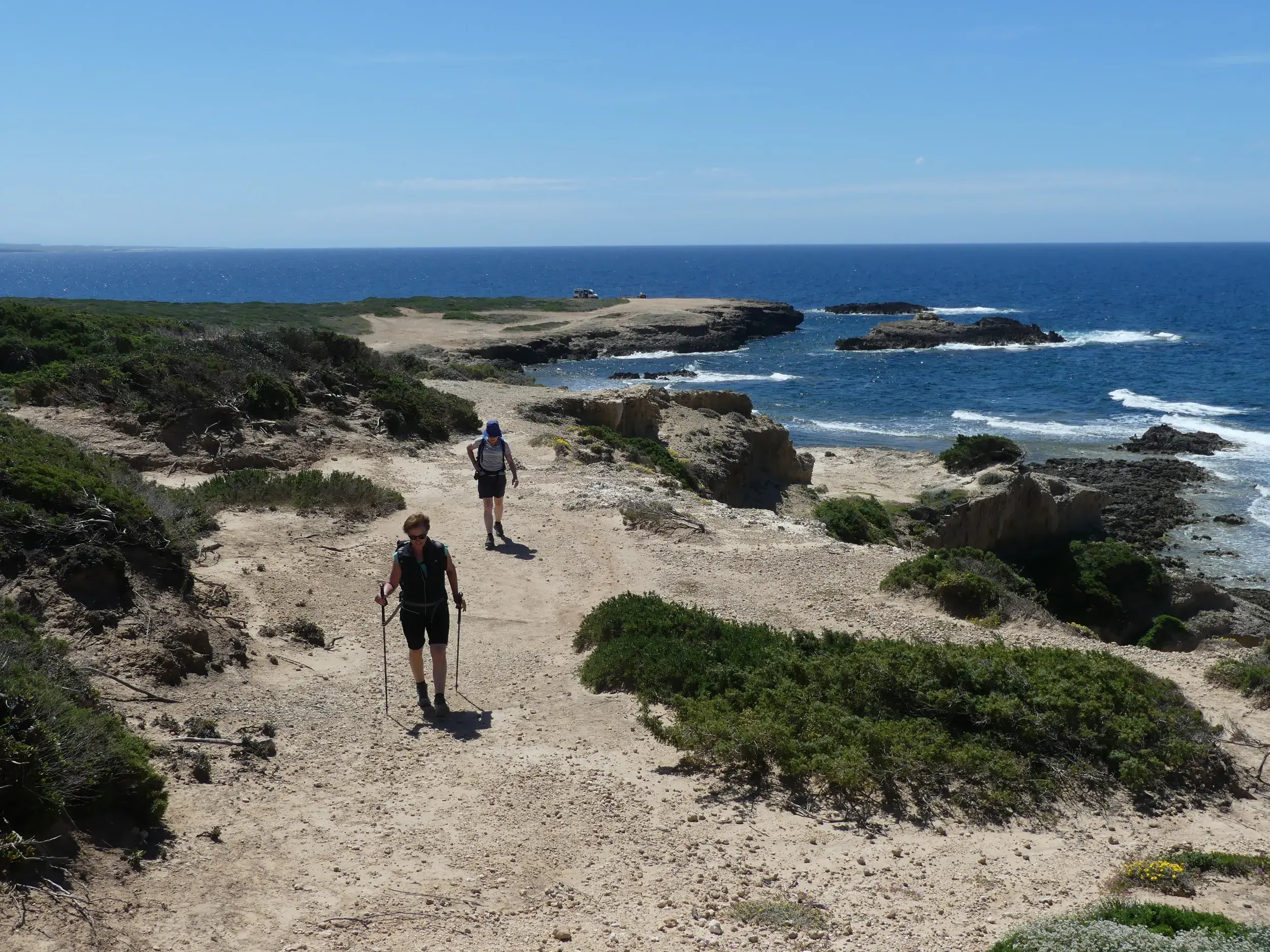 Zwei Wanderer auf einem sandigen Küstenpfad der Halbinsel Sinis mit Blick auf das felsige Ufer und das blaue Meer Sardiniens.