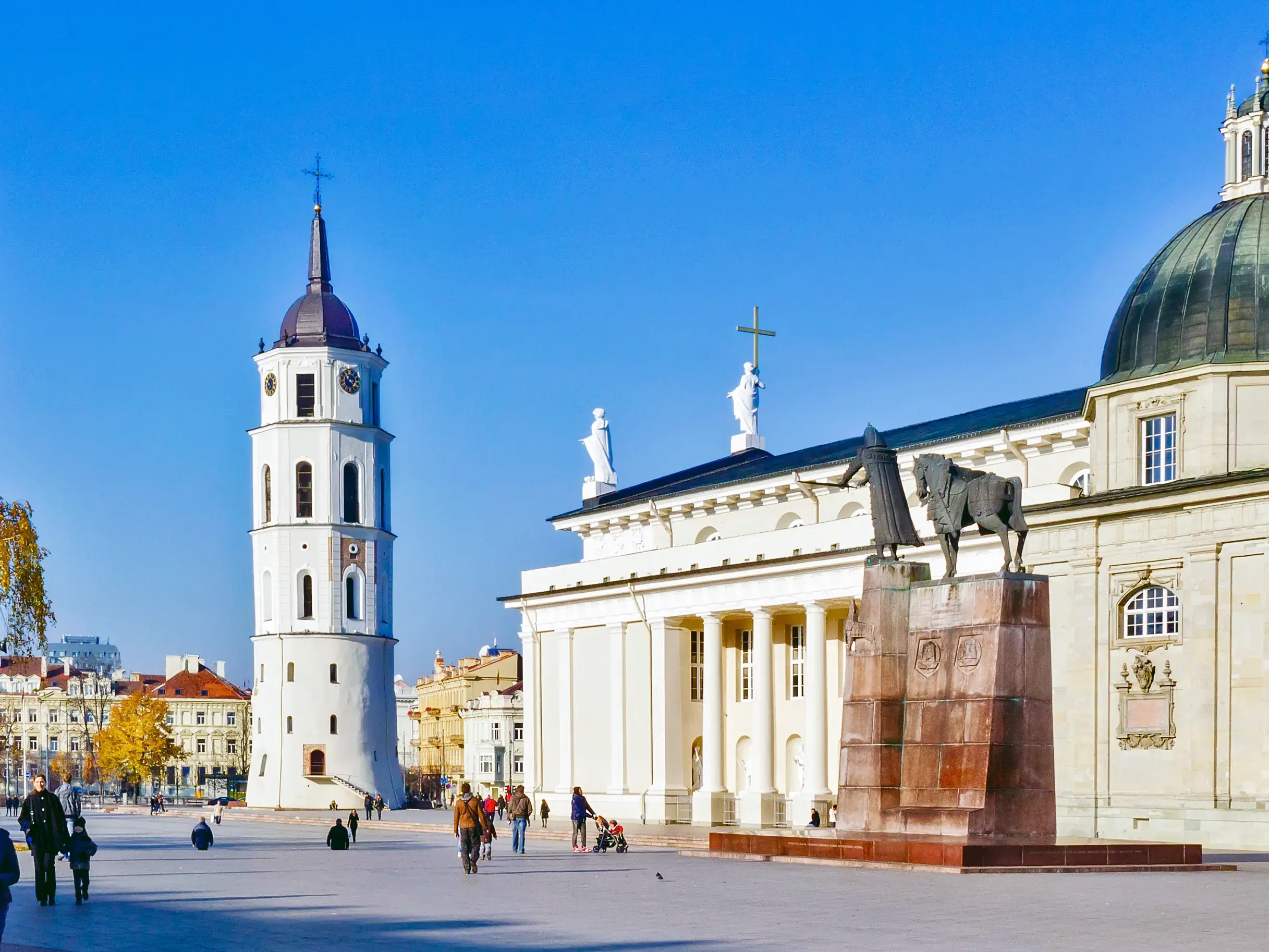 Die weißstrahlende Kathedrale von Vilnius und ihr Glockenturm an einem sonnigen Tag mit Passanten auf dem großen Vorplatz.
