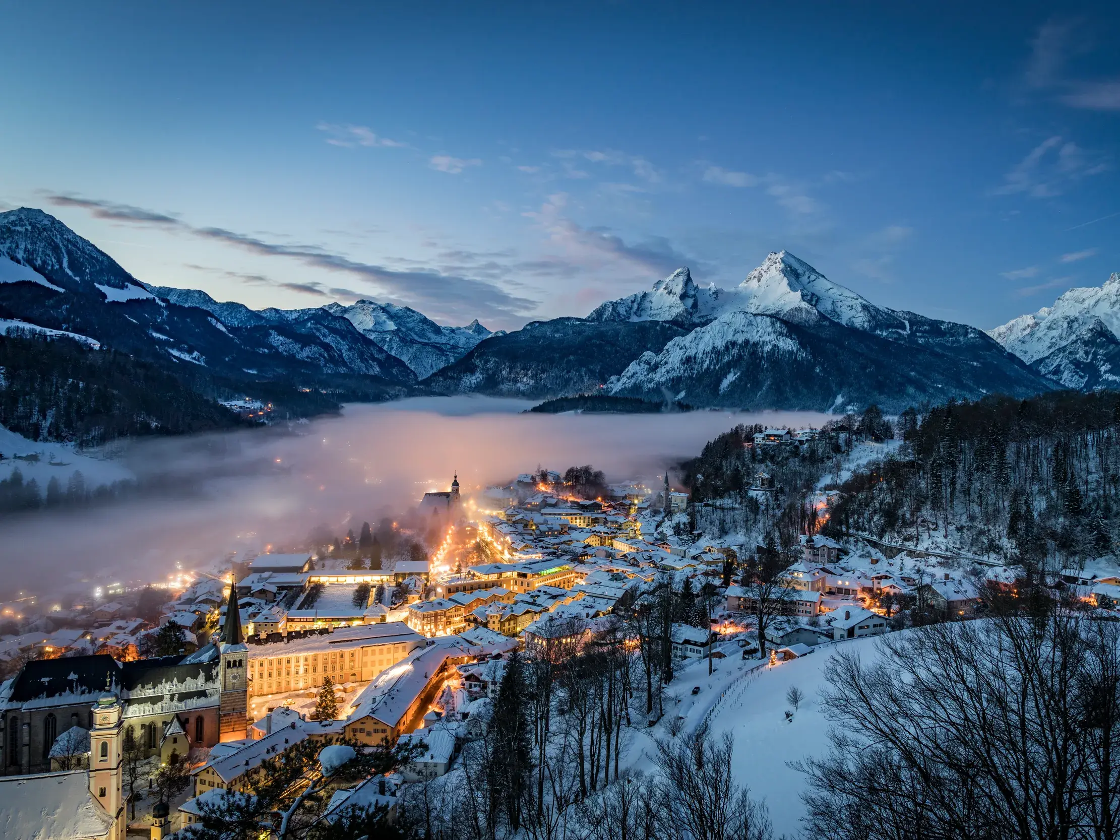 Abendlicher Blick über das verschneite Berchtesgaden, das im Talnebel leuchtet, überragt vom Watzmann-Massiv bei Dämmerung.