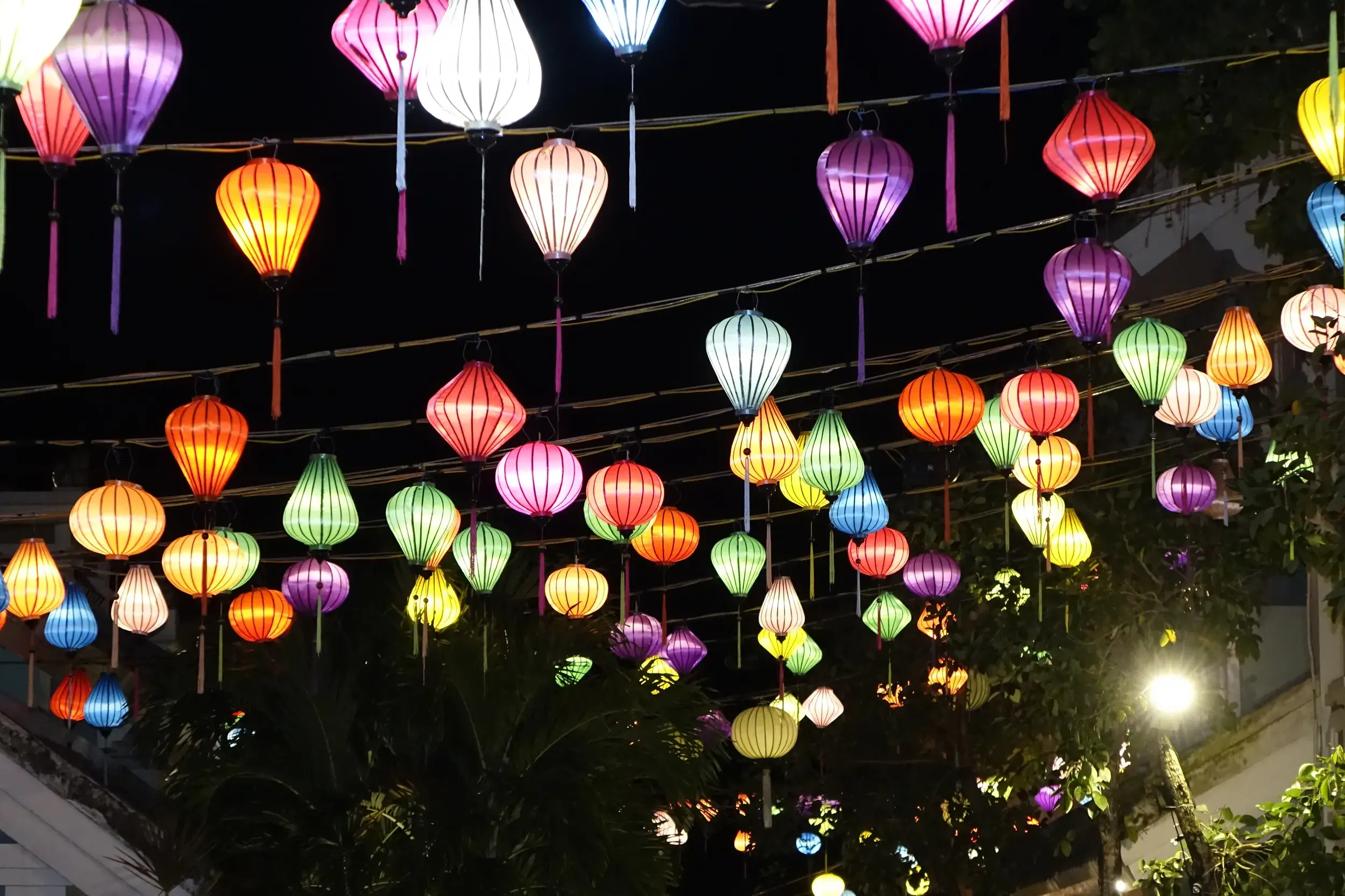 Bunte, leuchtende Seidenlampions in verschiedenen Farben und Formen hängen nachts über einer Straße in der Altstadt von Hoi An.