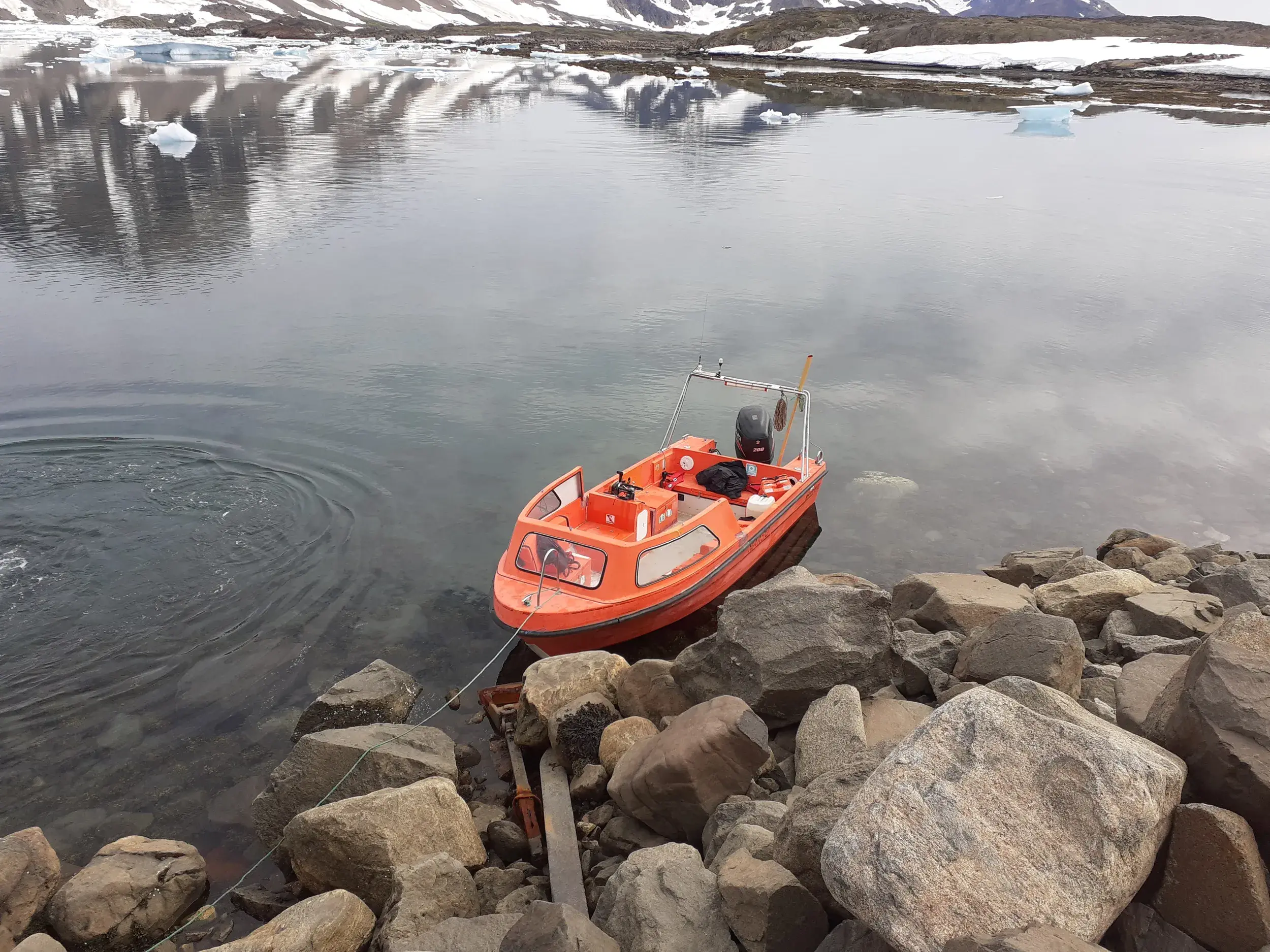 Oranges Motorboot am felsigen Ufer eines ruhigen Fjords, im Hintergrund spiegeln sich schneebedeckte Berge Grönlands.