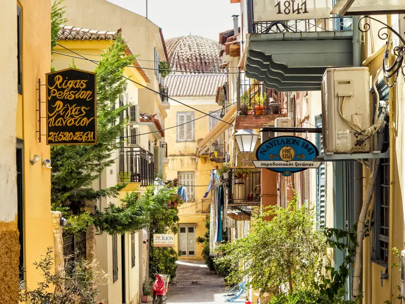 Sonnige Gasse in der historischen Altstadt von Nauplia mit blühenden Pflanzen, Balkonen und traditionellen Hotelschildern.