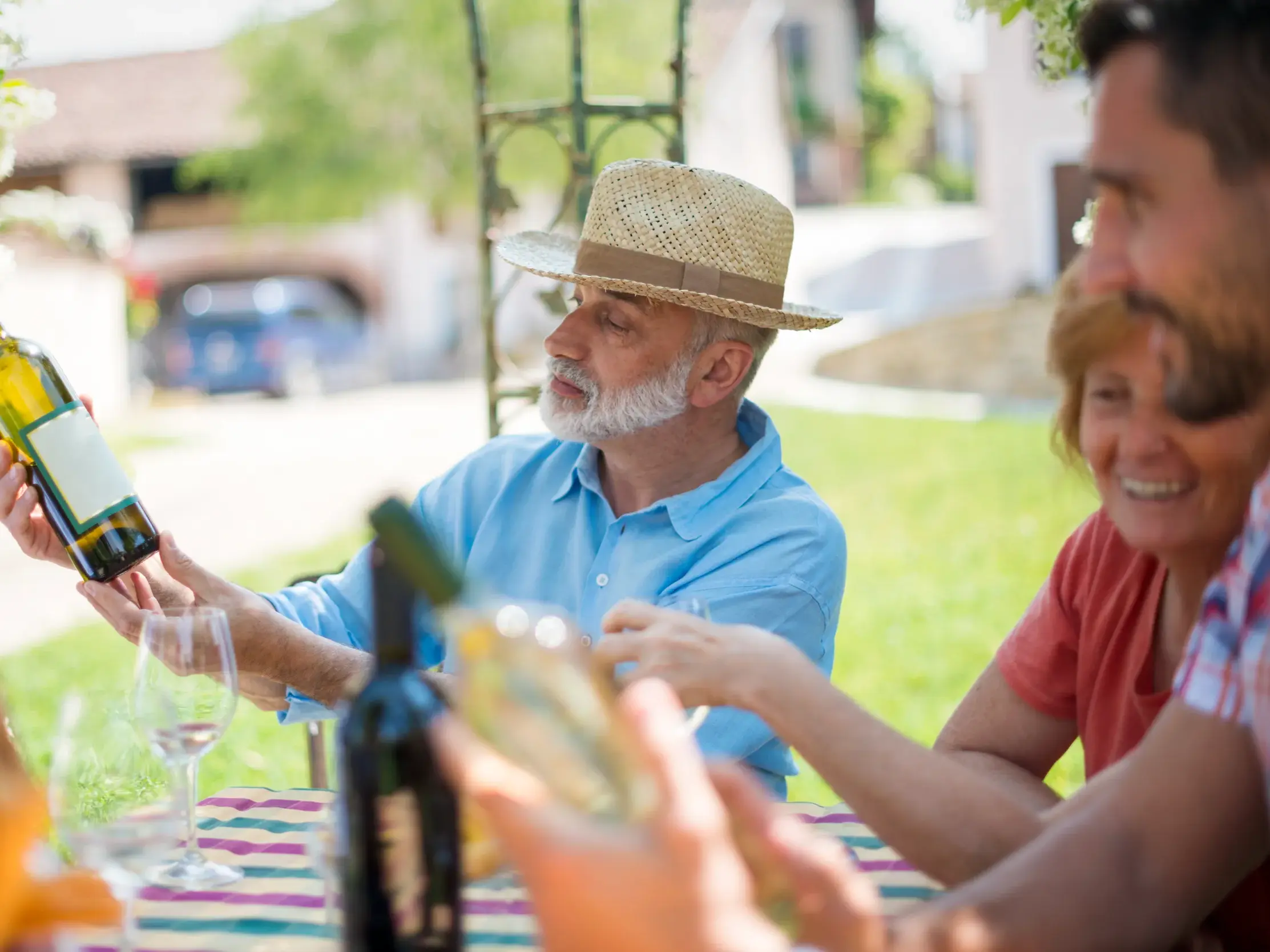 Auf einer Studienreise im Burgund begutachtet ein Senior mit Strohhut während einer Weinprobe im Freien eine Flasche Weißwein