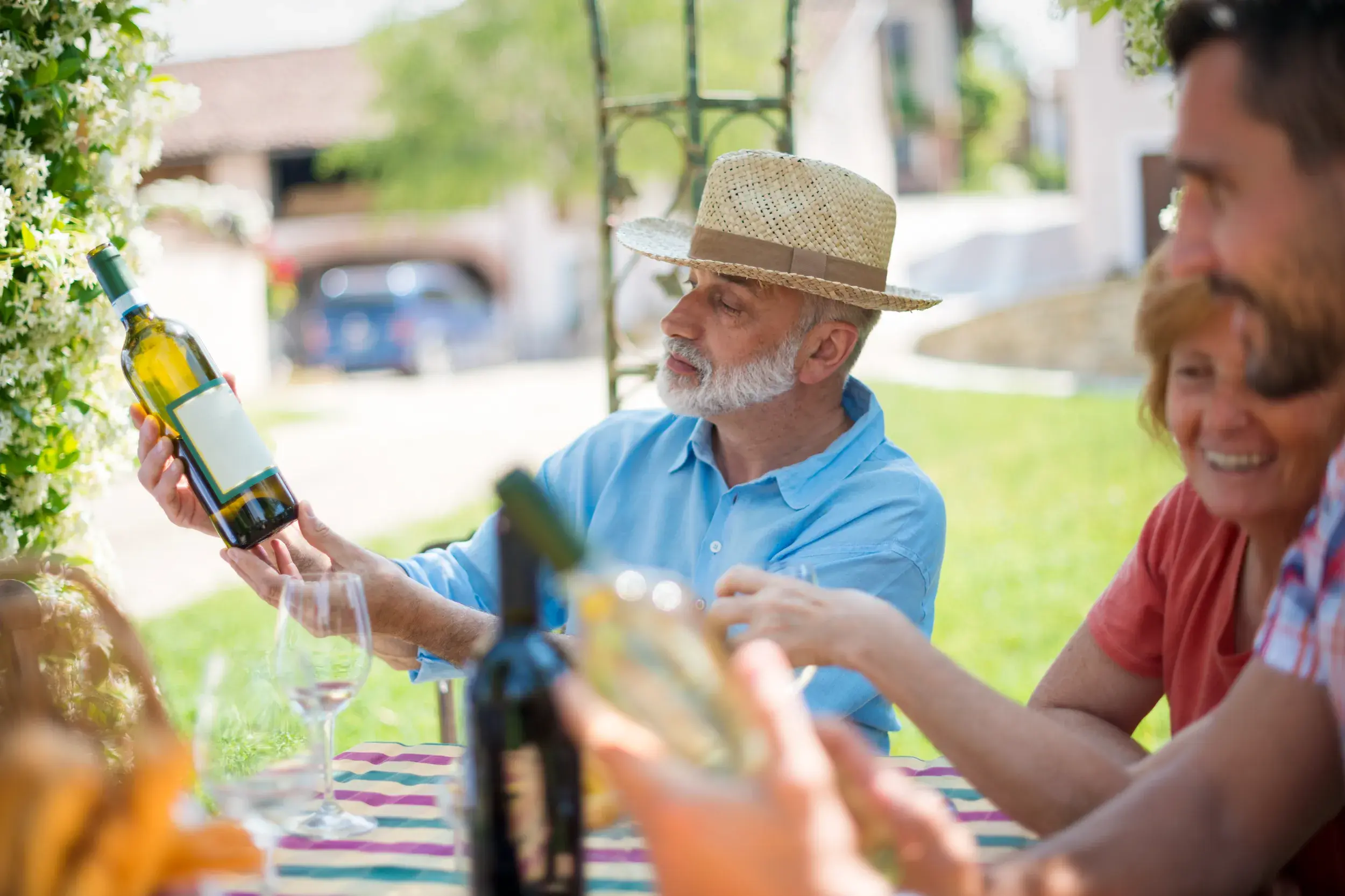 Auf einer Studienreise im Burgund begutachtet ein Senior mit Strohhut während einer Weinprobe im Freien eine Flasche Weißwein