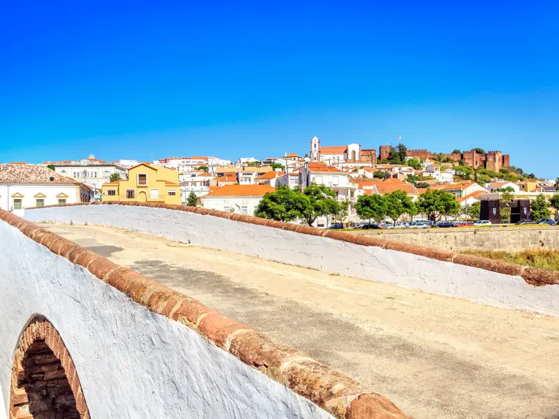 Blick von einer historischen Brücke auf die Stadt Silves mit der mächtigen Maurenburg unter blauem Himmel an der Algarve.