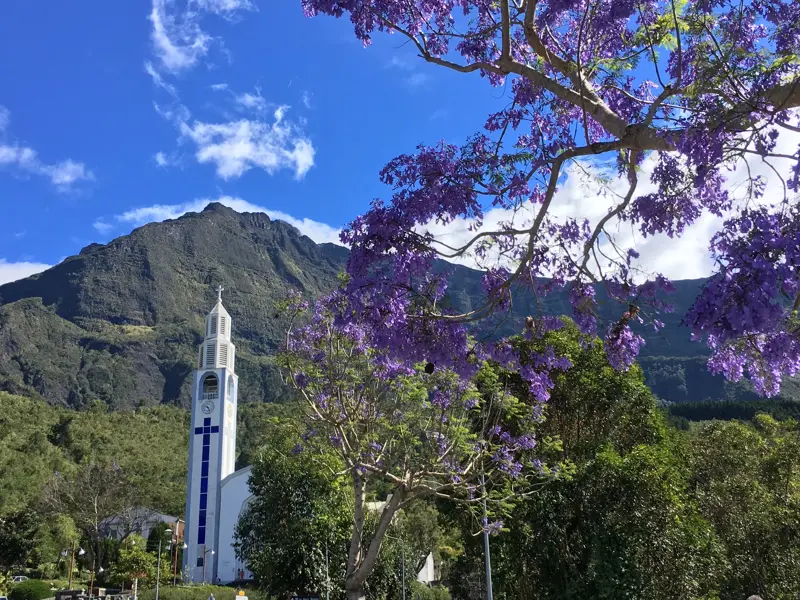Die weiße Kirche von Cilaos im Bergkessel Cirque de Cilaos, umrahmt von den lila Blüten eines Jacaranda-Baums.