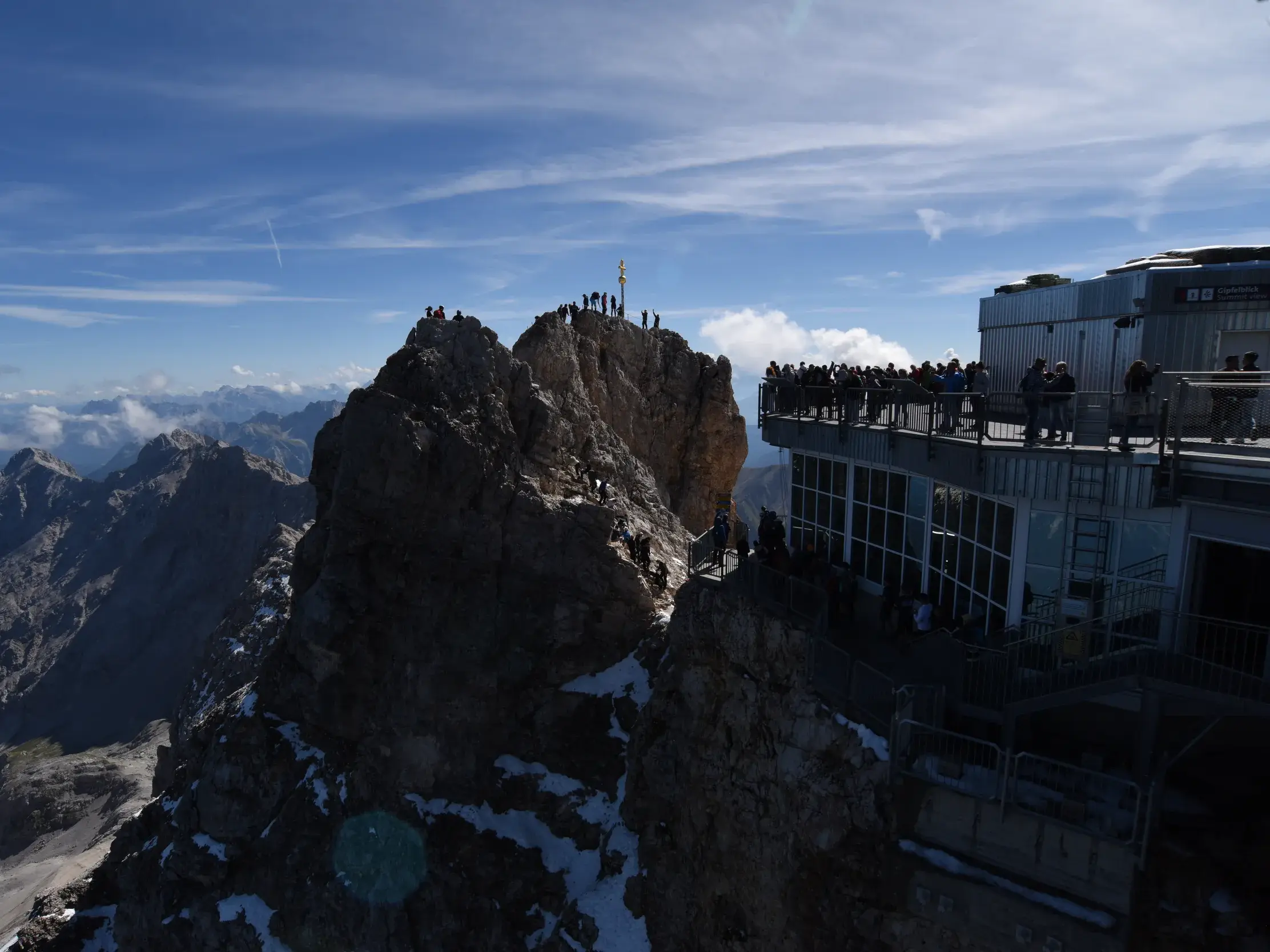 Bergsteiger auf dem Felsgipfel der Zugspitze neben der Besucherplattform mit Panoramablick über die Alpen bei Sonnenschein.