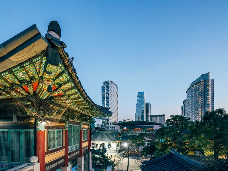 Verziertes Dach des Jogyesa-Tempels in Seoul bei Dämmerung mit der beleuchteten Skyline moderner Hochhäuser im Hintergrund.