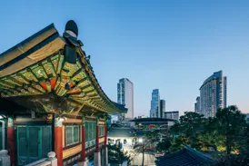Verziertes Dach des Jogyesa-Tempels in Seoul bei Dämmerung mit der beleuchteten Skyline moderner Hochhäuser im Hintergrund.