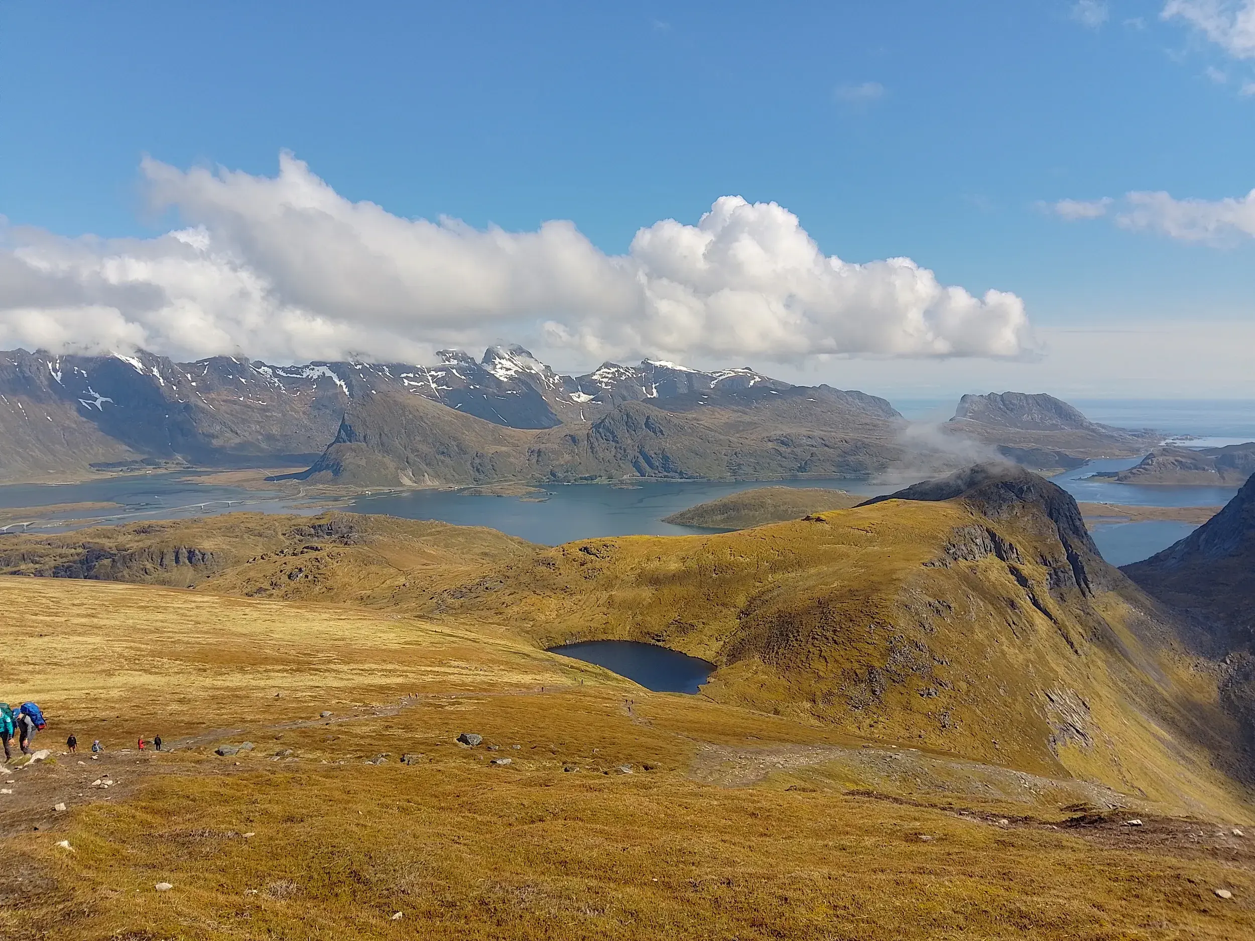 Eine kleine Wandergruppe auf einem weiten, goldenen Hochplateau mit Blick auf die zerklüftete Fjordlandschaft der Lofoten.