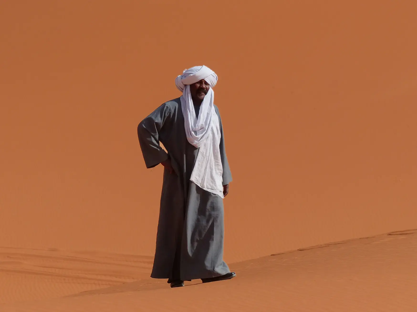 Ein Tuareg-Begleiter in grauer Robe und weißem Turban steht auf einer leuchtend orangen Sanddüne im Erg Admer, Algerien.