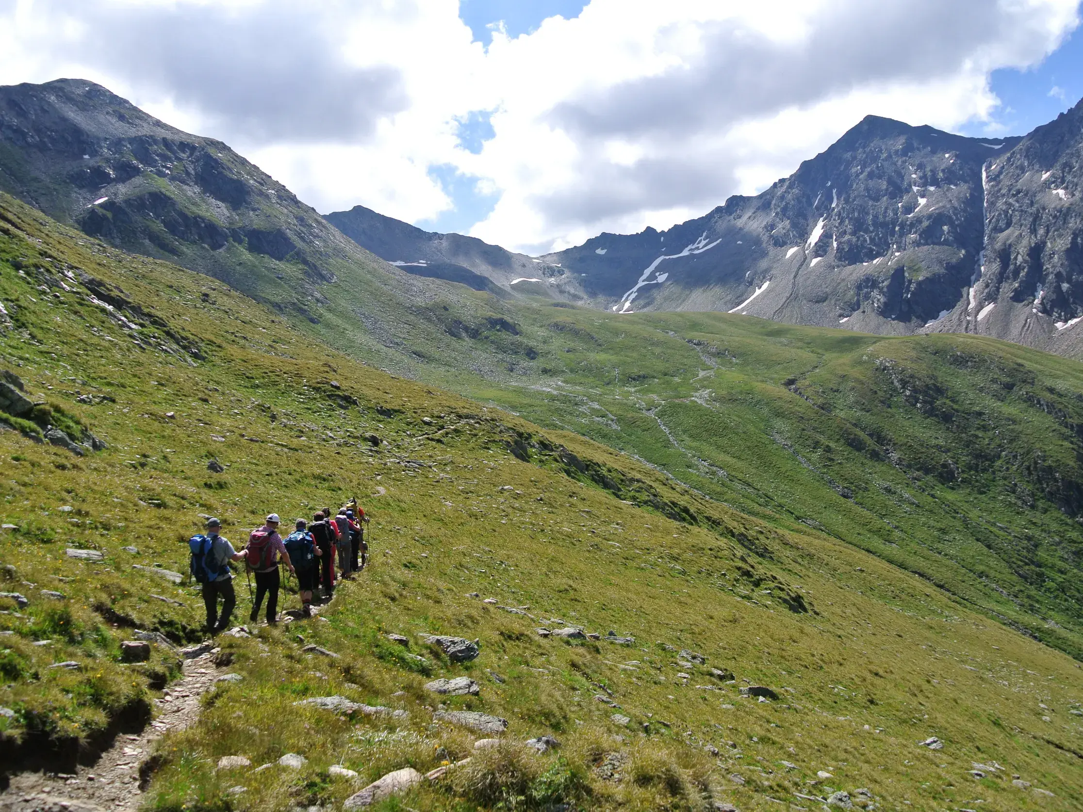 Eine Wandergruppe mit Rucksäcken auf einem schmalen Pfad an einem grünen Hang im hochalpinen Nationalpark Hohe Tauern.