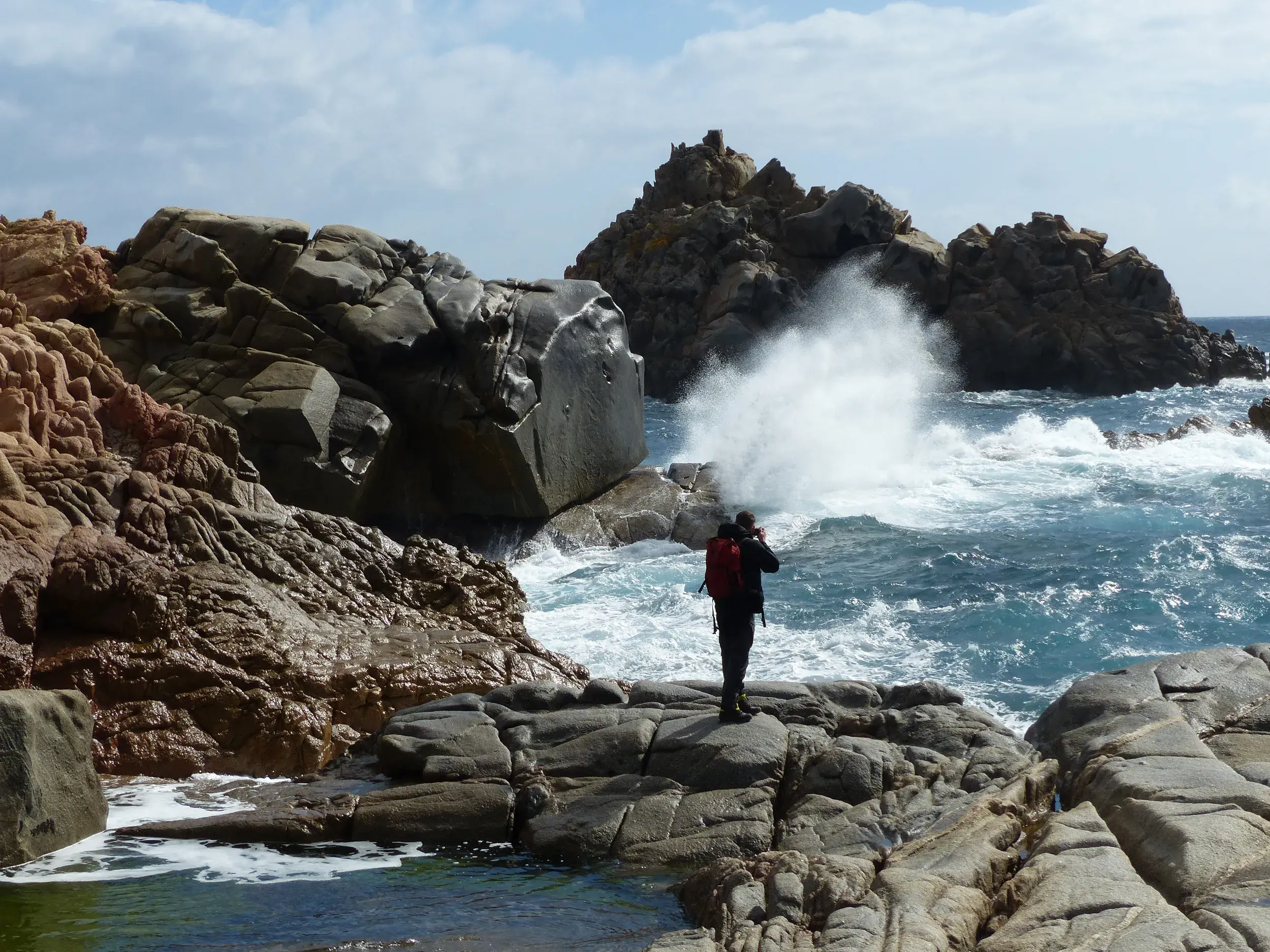 Ein Wanderer fotografiert auf einer Küstenwanderung auf Sardinien eine große Welle, die an der roten Felsküste bricht.