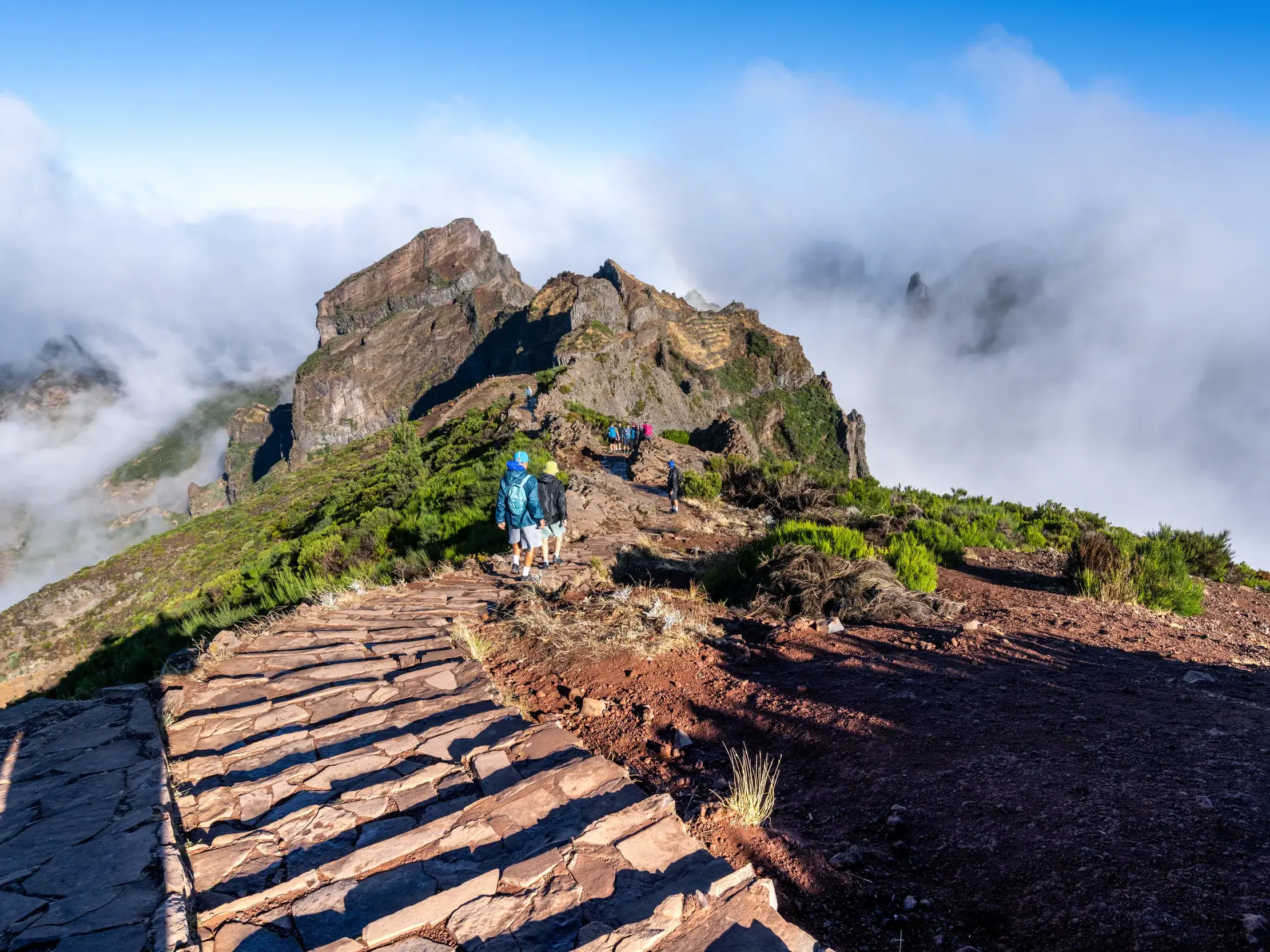 Eine kleine Wandergruppe erkundet auf einem gesicherten Pfad die schroffen Gipfel am Pico do Arieiro auf Madeira über den Wolken.