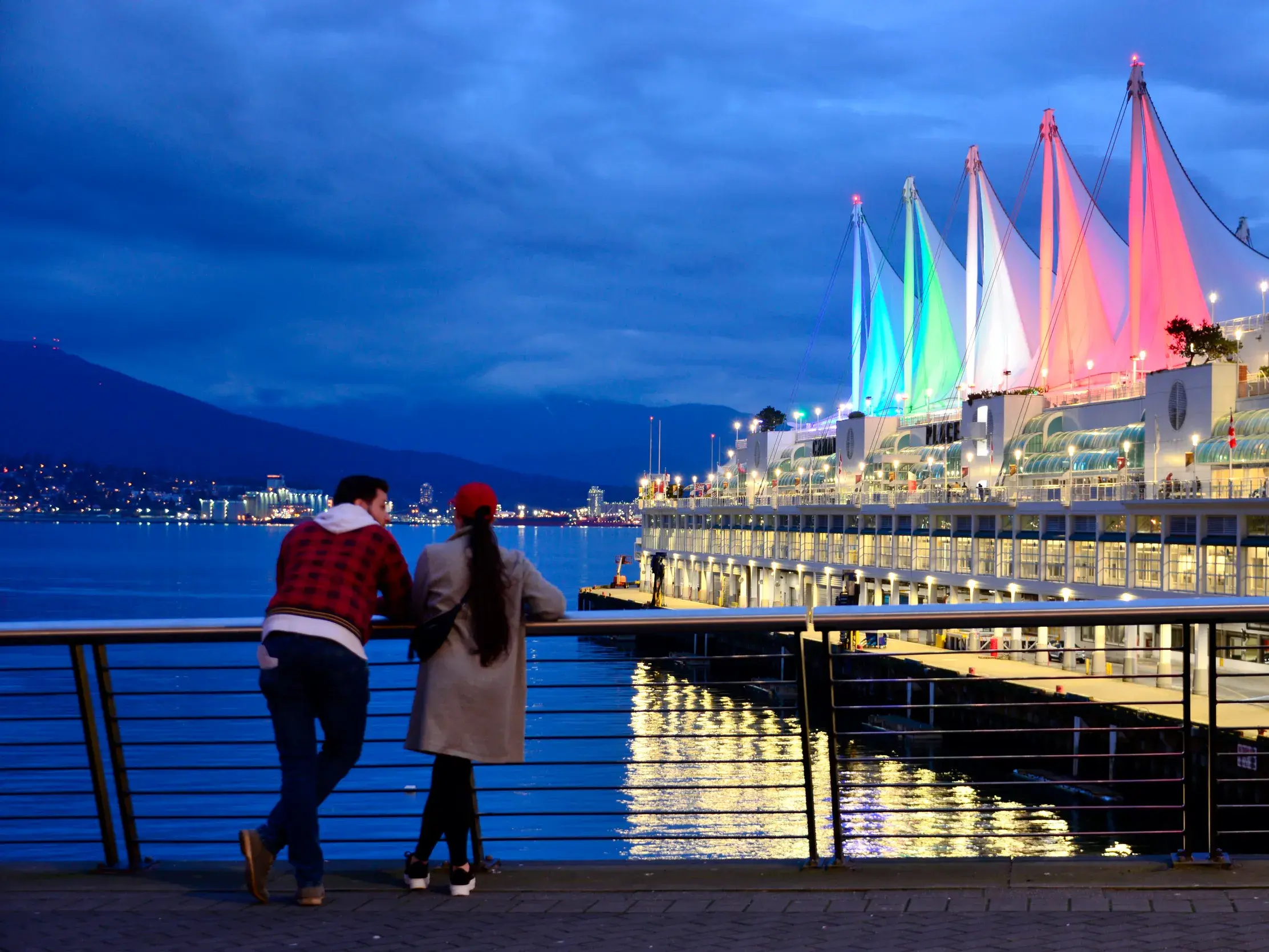 Zwei junge Reisende genießen bei Dämmerung den Blick auf das farbenfroh beleuchtete Canada Place in Vancouver.