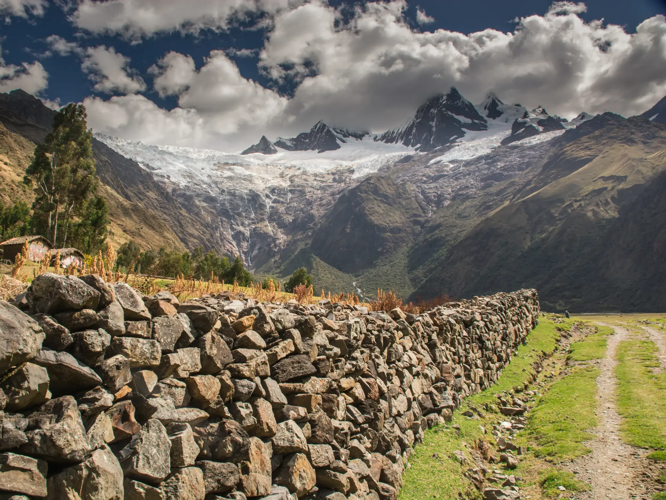 Eine rustikale Steinmauer führt einen Wanderweg entlang durch die peruanische Cordillera Blanca mit Blick auf massive Gletscher.