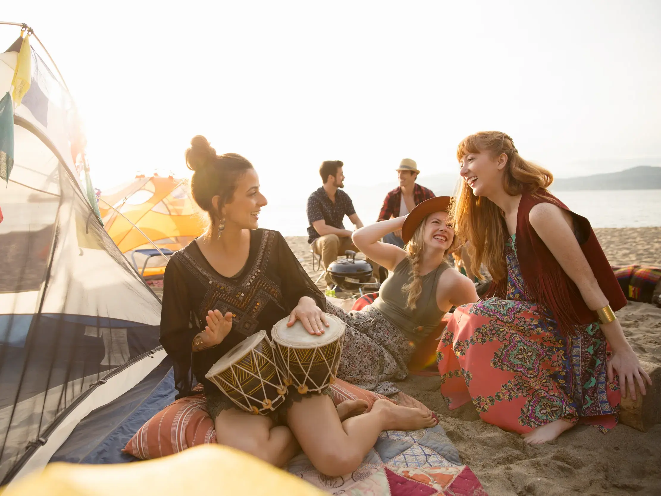 Junge Reisegruppe entspannt am Strand nahe Neapel, eine Frau spielt Trommel und ihre Freunde lachen im Sonnenuntergang.