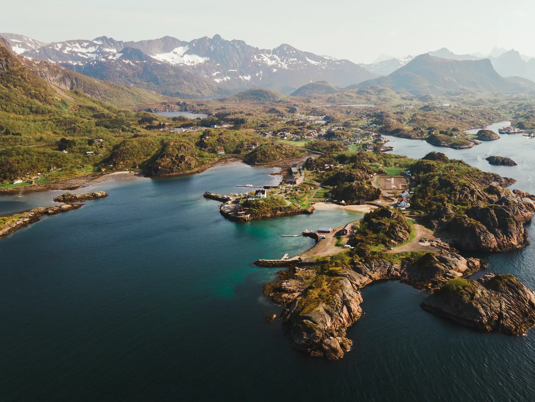 Weitblick über die grüne, zerklüftete Inselwelt der Lofoten mit tiefblauen Fjorden und schneebedeckten Bergen im Hintergrund.