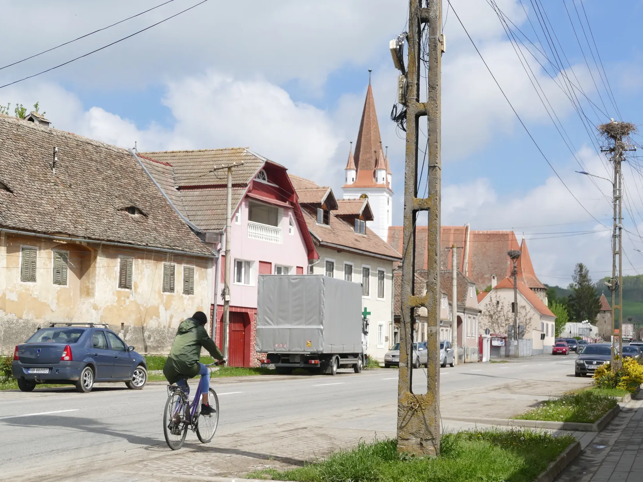 Sonnige Dorfstraße in Siebenbürgen mit traditionellen Häusern mit Augenfach-Fenstern, Storchennestern und einer Radfahrerin.