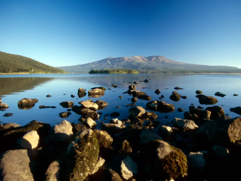 Felsiges Ufer am Lake Taupo mit klarem Wasser und Blick auf die Vulkane des Tongariro Nationalparks an einem sonnigen Tag.