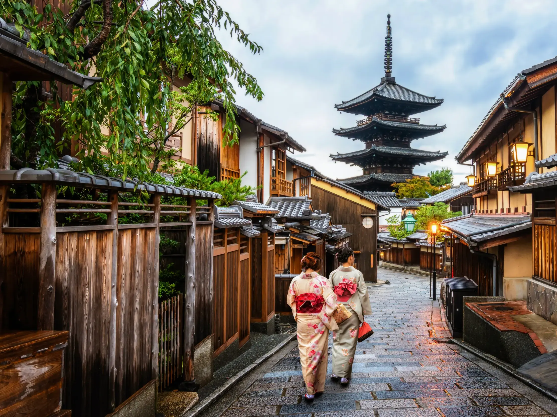 Zwei Frauen in Kimonos auf einer Gasse im Gion-Viertel in Kyoto, gesäumt von Holzhäusern, mit Blick auf die Yasaka-Pagode.