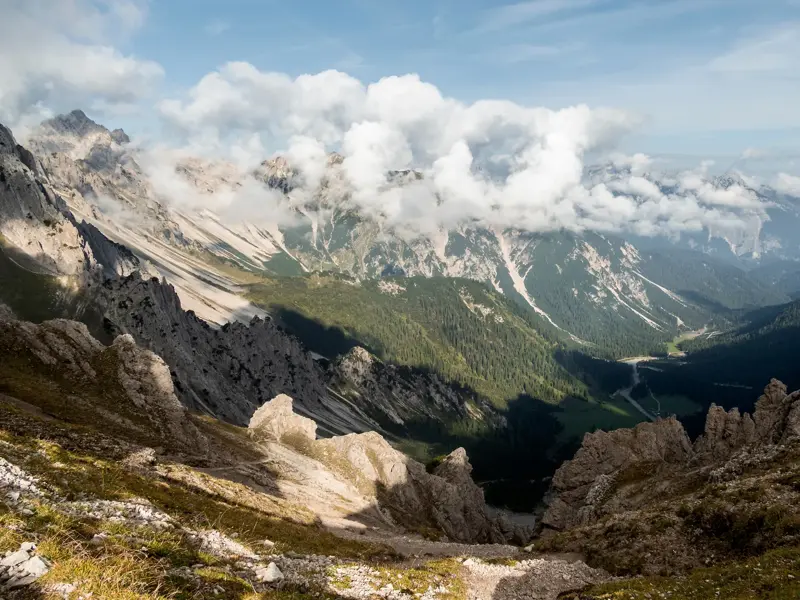 Grandiose Aussicht vom Gipfel beim Trekking im Karwendelgebirge auf ein tiefes, bewaldetes Tal und wolkenverhangene Alpengipfel.