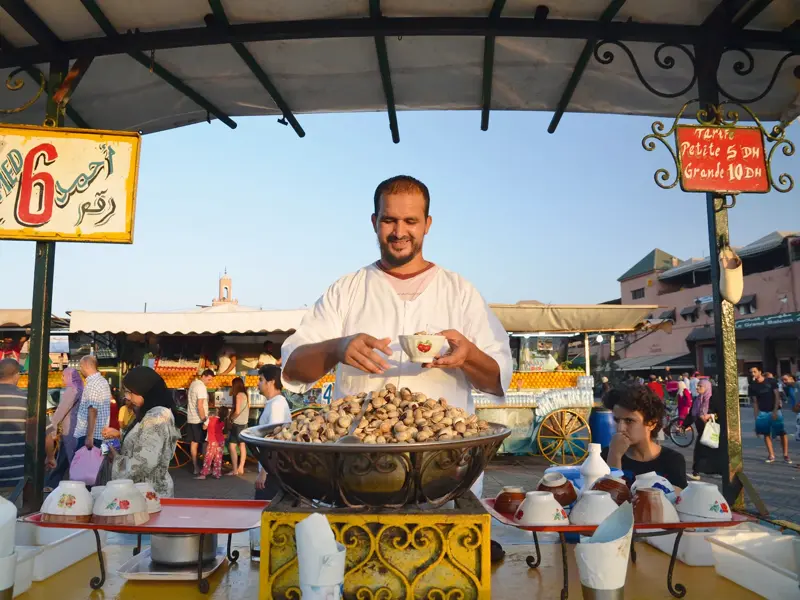 Ein freundlicher Verkäufer serviert an seinem Stand auf dem Djemaa el Fna in Marrakesch eine Portion gekochter Schnecken.