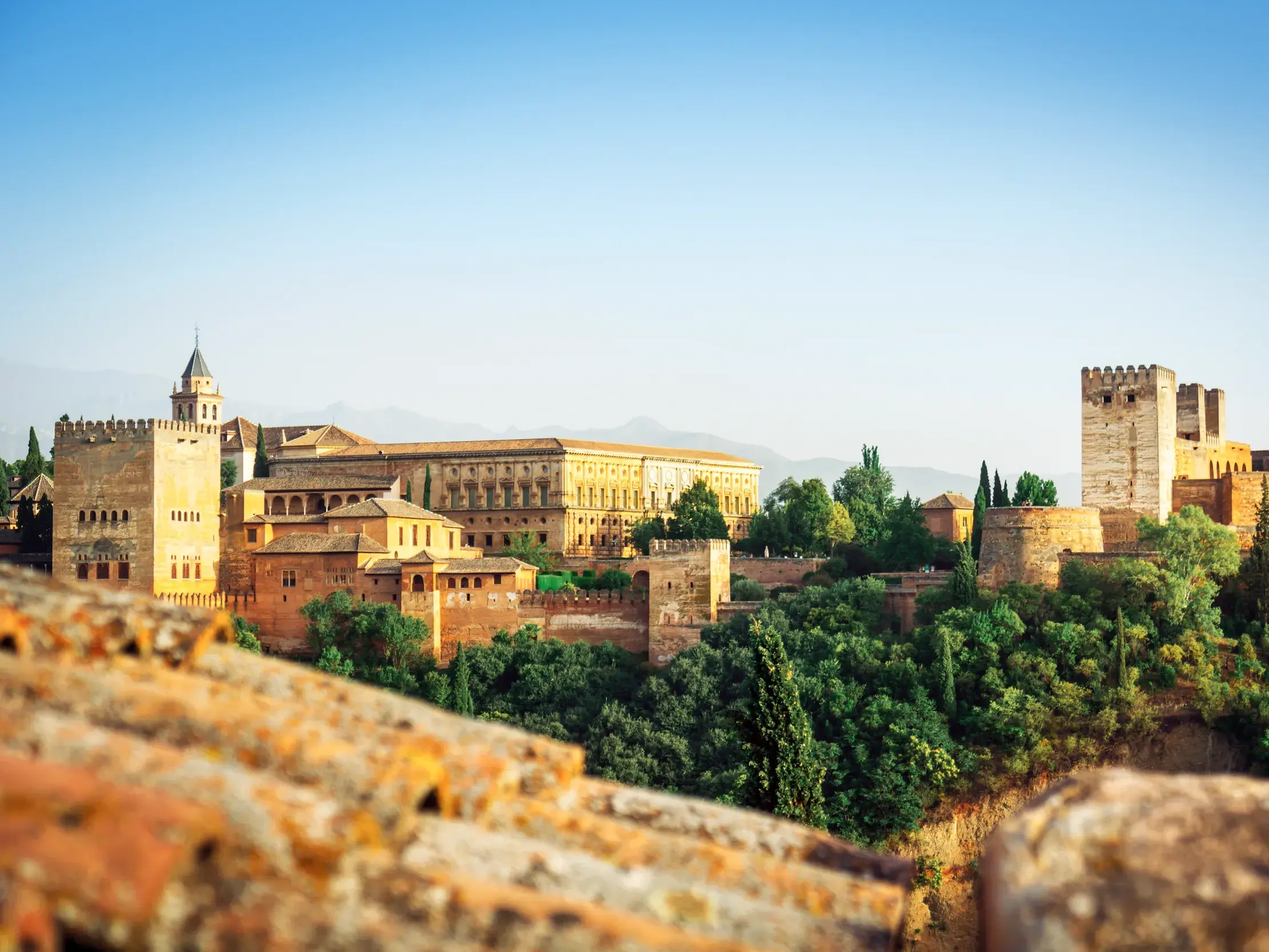 Blick über die Ziegeldächer des Albaicín auf die Festungsanlage der Alhambra in Granada vor der Silhouette der Sierra Nevada.