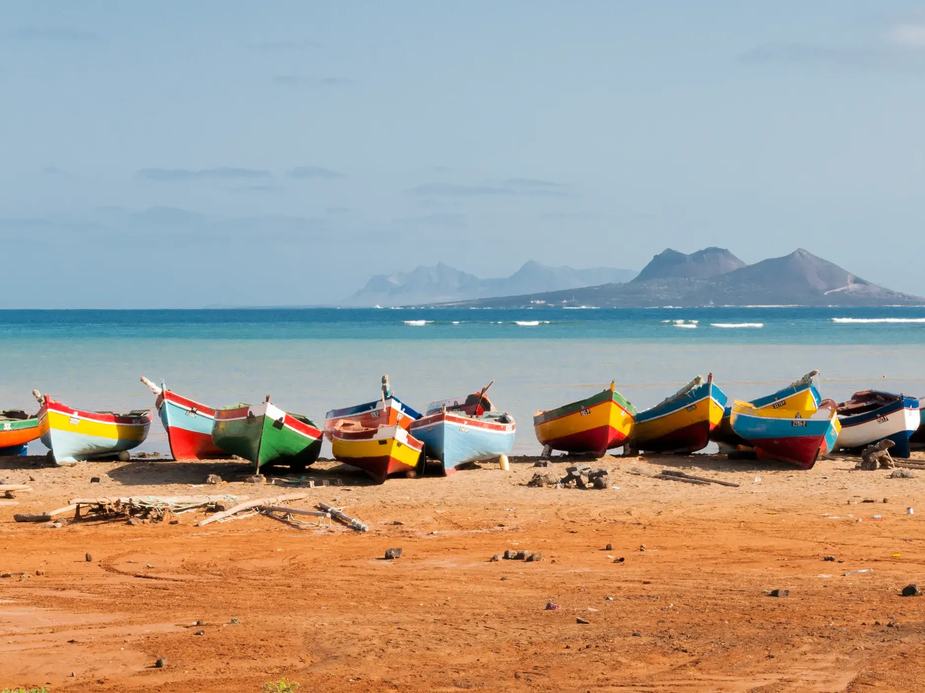 Zahlreiche bunte Fischerboote liegen an einem Sandstrand auf den Kapverden, im Hintergrund das türkise Meer und eine Bergkette.