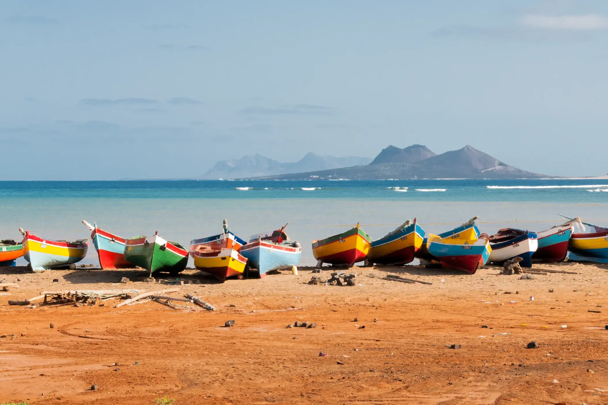 Zahlreiche bunte Fischerboote liegen an einem Sandstrand auf den Kapverden, im Hintergrund das türkise Meer und eine Bergkette.