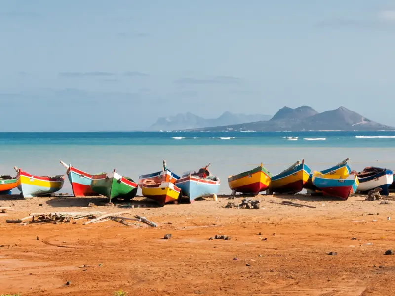 Zahlreiche bunte Fischerboote liegen an einem Sandstrand auf den Kapverden, im Hintergrund das türkise Meer und eine Bergkette.