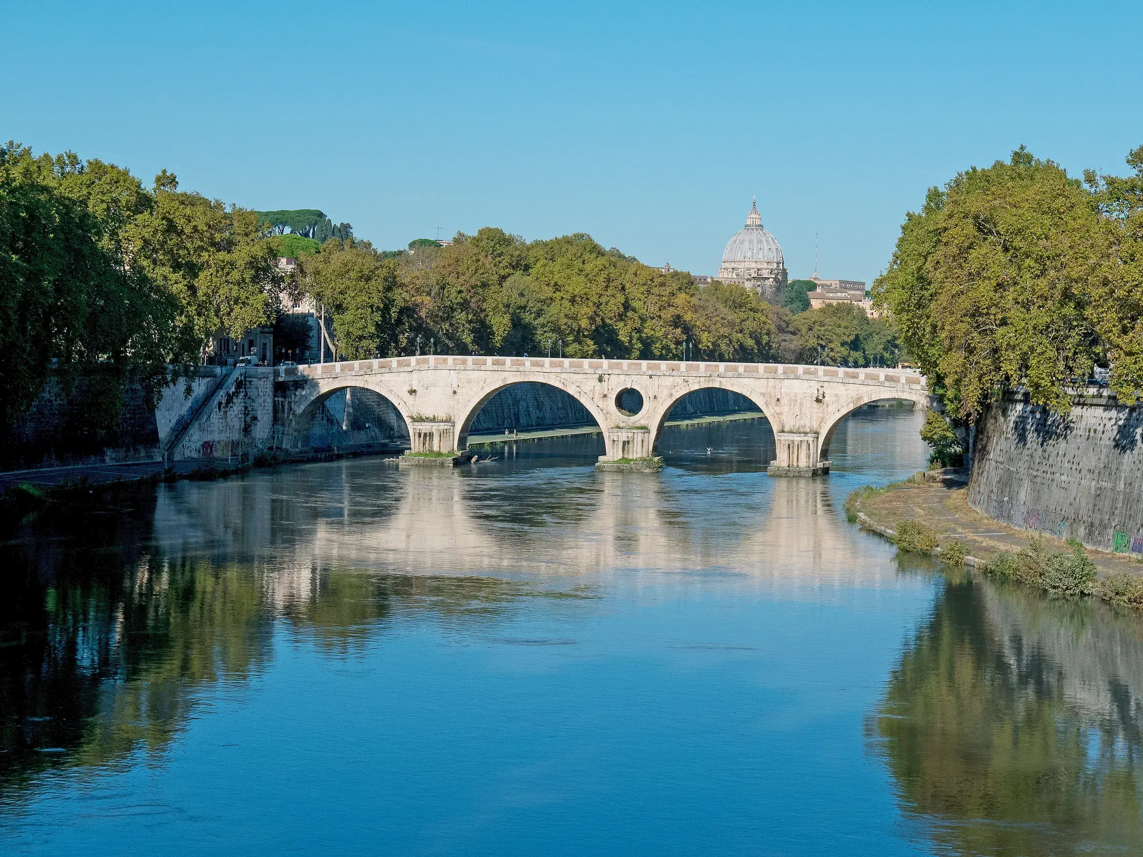 Sonniger Blick über den Tiber auf die Ponte Sisto in Rom, eine historische Steinbrücke mit dem Petersdom im Hintergrund.