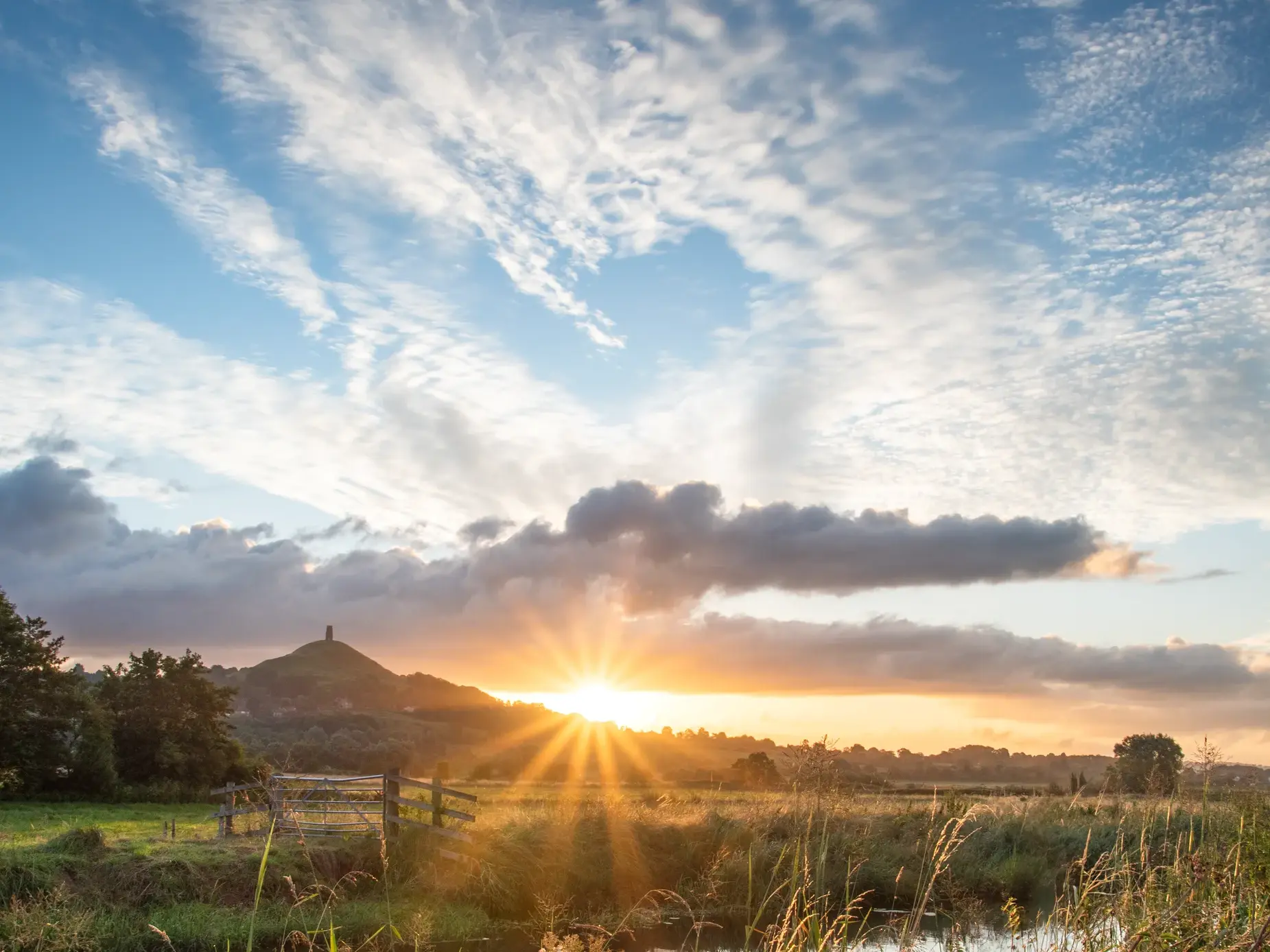 Die aufgehende Sonne taucht Glastonbury in goldenes Licht, Gräser am Flussufer leuchten.