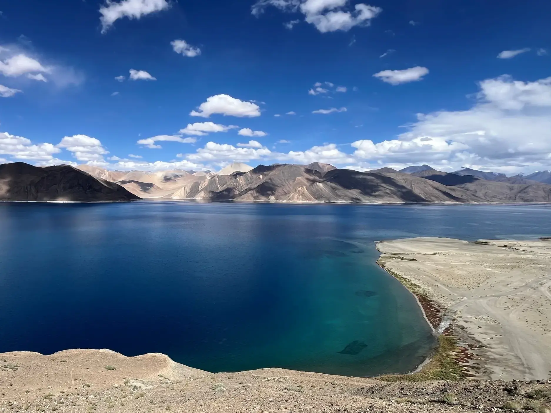 Panoramablick auf den tiefblauen Pangong See in Ladakh, umgeben von kargen Bergen des Himalaya unter blauem Himmel mit Wolken.