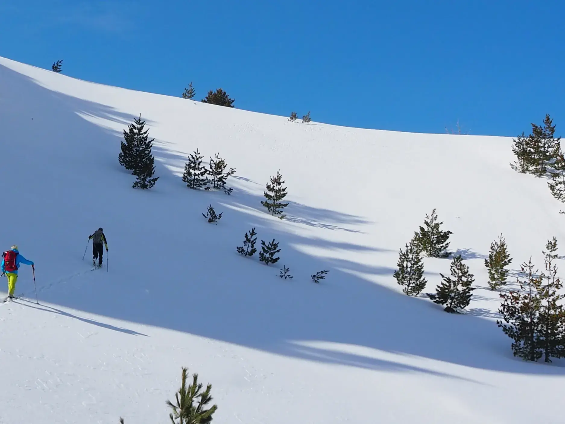 Zwei Skitourengeher beim Aufstieg auf einem unberührten Schneehang im Balkan bei strahlend blauem Himmel.