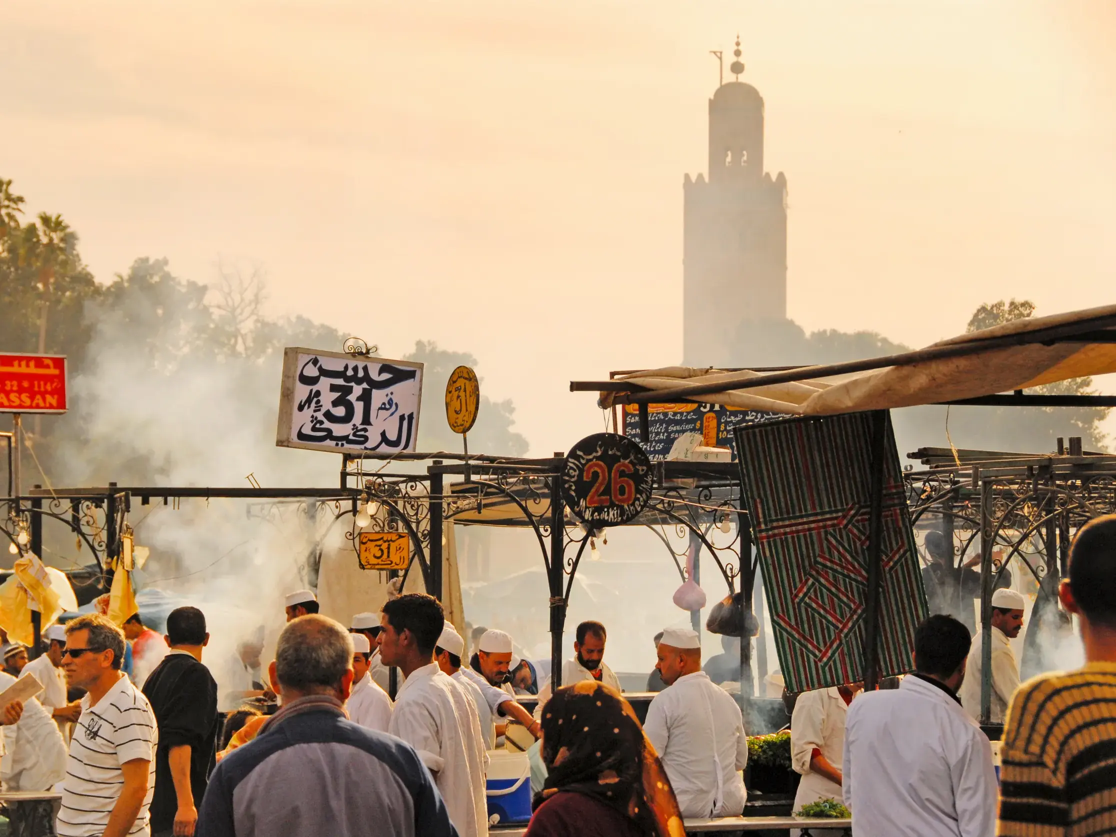 Lebhafter Abend auf dem Djemma el Fna in Marrakesch mit Garküchen, Rauchschwaden und der Koutoubia-Moschee im Hintergrund.
