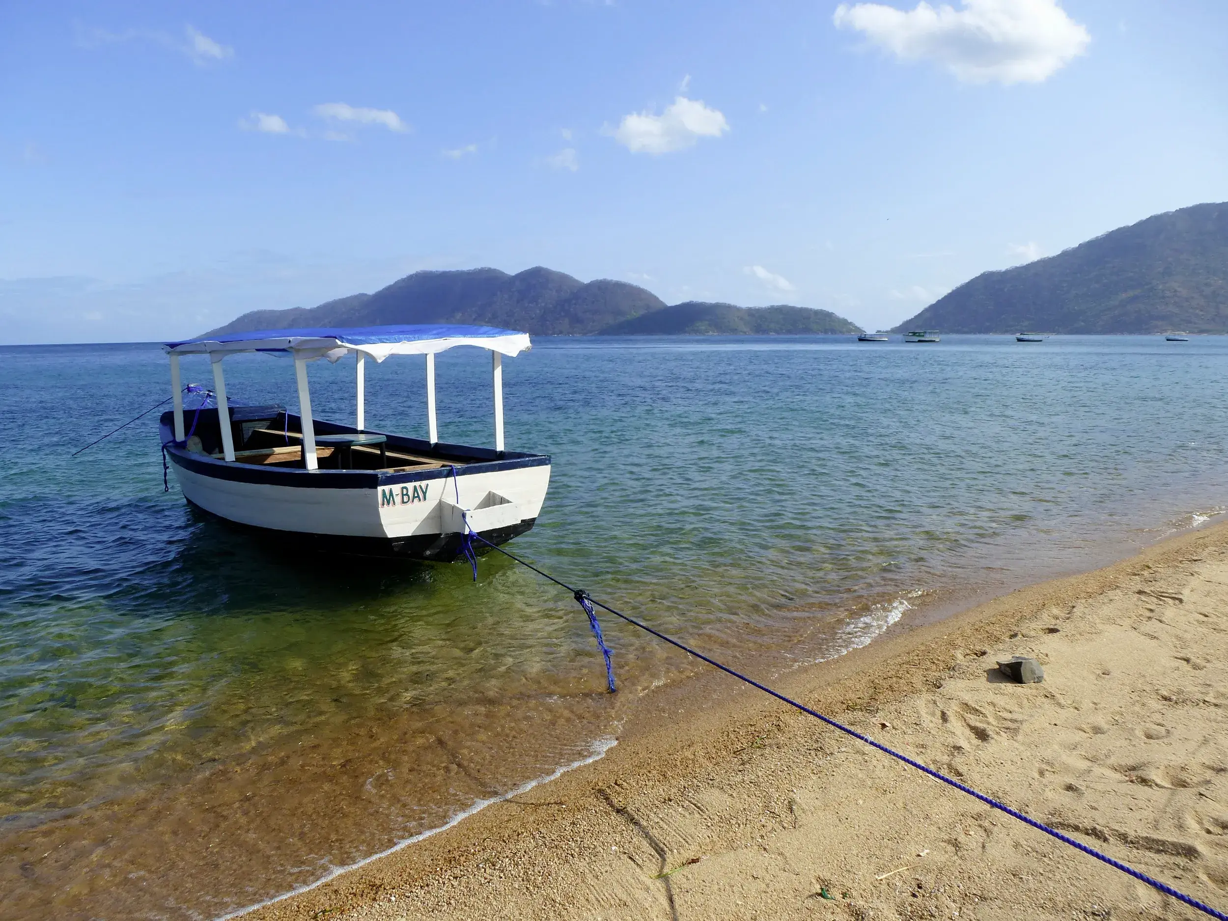 Ein weißes Boot mit blauem Dach liegt im klaren Wasser am Sandstrand des Malawi-Sees vor der Kulisse grüner Hügel.