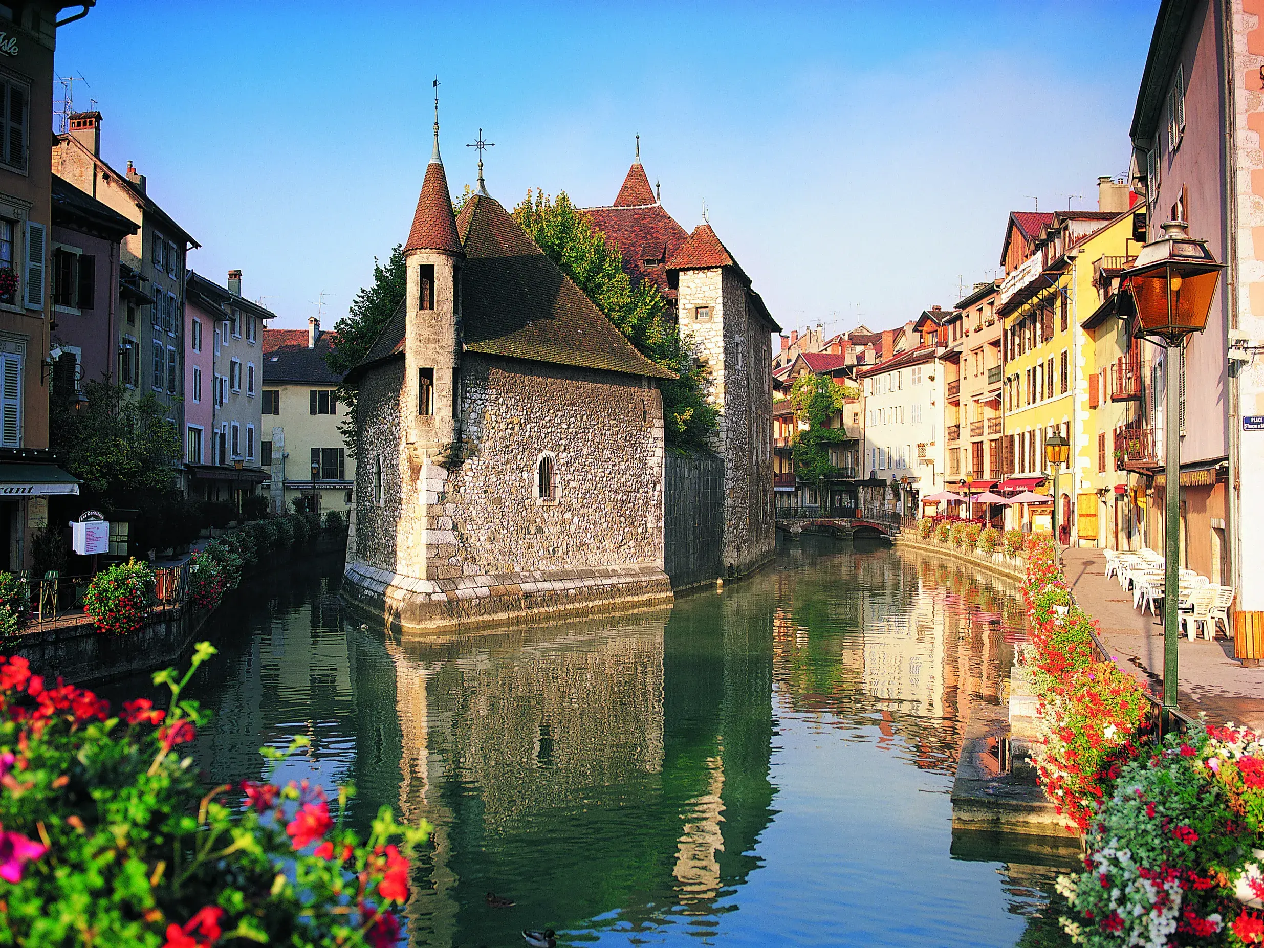 Das historische Palais de l'Isle in Annecy, umflossen von einem Kanal, mit bunten Altstadthäusern bei strahlend blauem Himmel.