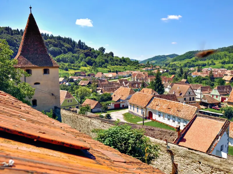 Idyllischer Blick über die roten Ziegeldächer eines Dorfes in Siebenbürgen mit Wehrturm vor den grünen Karpaten.